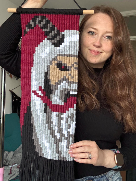 Woman holding a pixelated macrame wall hanging with a design of Krampus.