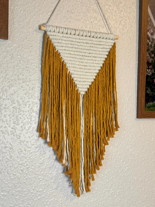 Woven wall hanging with mustard yellow and white fringes on a textured wall.