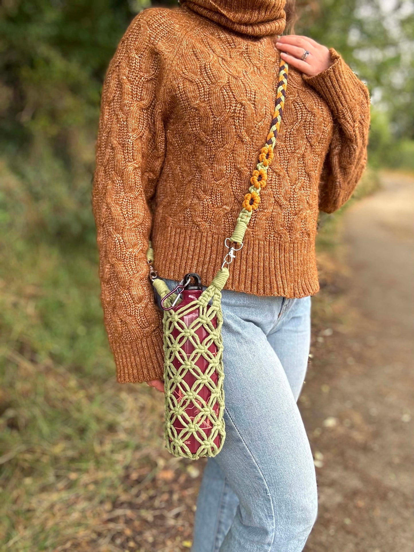 A person outdoors wearing a brown cable-knit turtleneck sweater and light blue jeans, carrying a woven crossbody bag with a yellow and green patterned strap with sunflowers