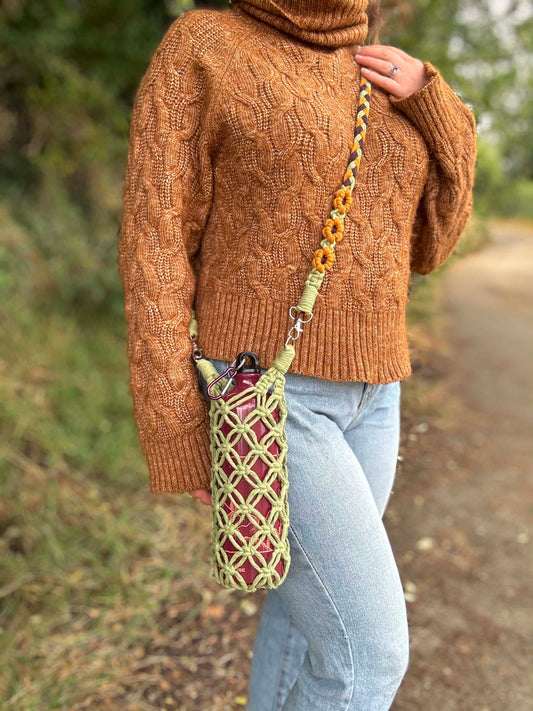 A person outdoors wearing a brown cable-knit turtleneck sweater and light blue jeans, carrying a woven crossbody bag with a yellow and green patterned strap with sunflowers