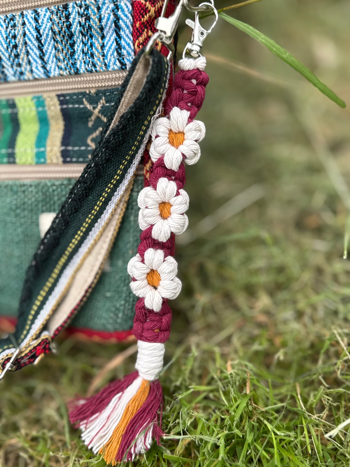 A close-up of a colorful woven and embroidered bag charm with white daisy flowers, a maroon braided strap, and a tassel, hanging from a zipper on an outdoor grassy surface.