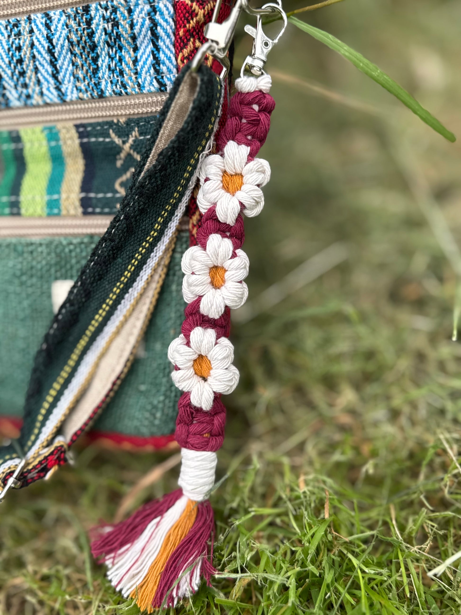 A close-up of a colorful woven and embroidered bag charm with white daisy flowers, a maroon braided strap, and a tassel, hanging from a zipper on an outdoor grassy surface.