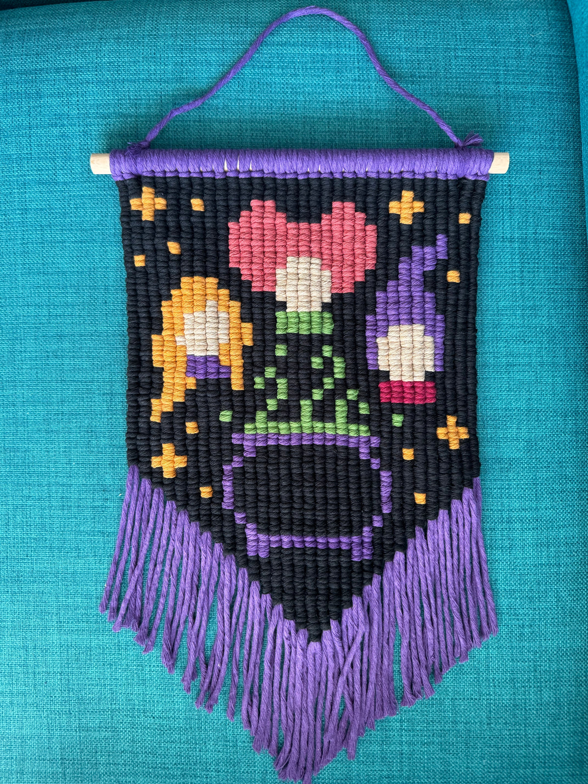 Macrame wall hanging featuring a pixelised version of the Sanderson sisters from Hocus Pocus and a bubbling cauldron, with a black background and a purple fringe.