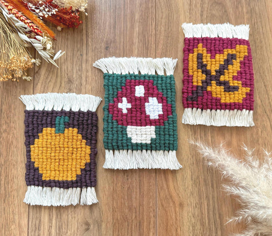 Three macrame coasters with autumn-themed designs on a wooden surface.