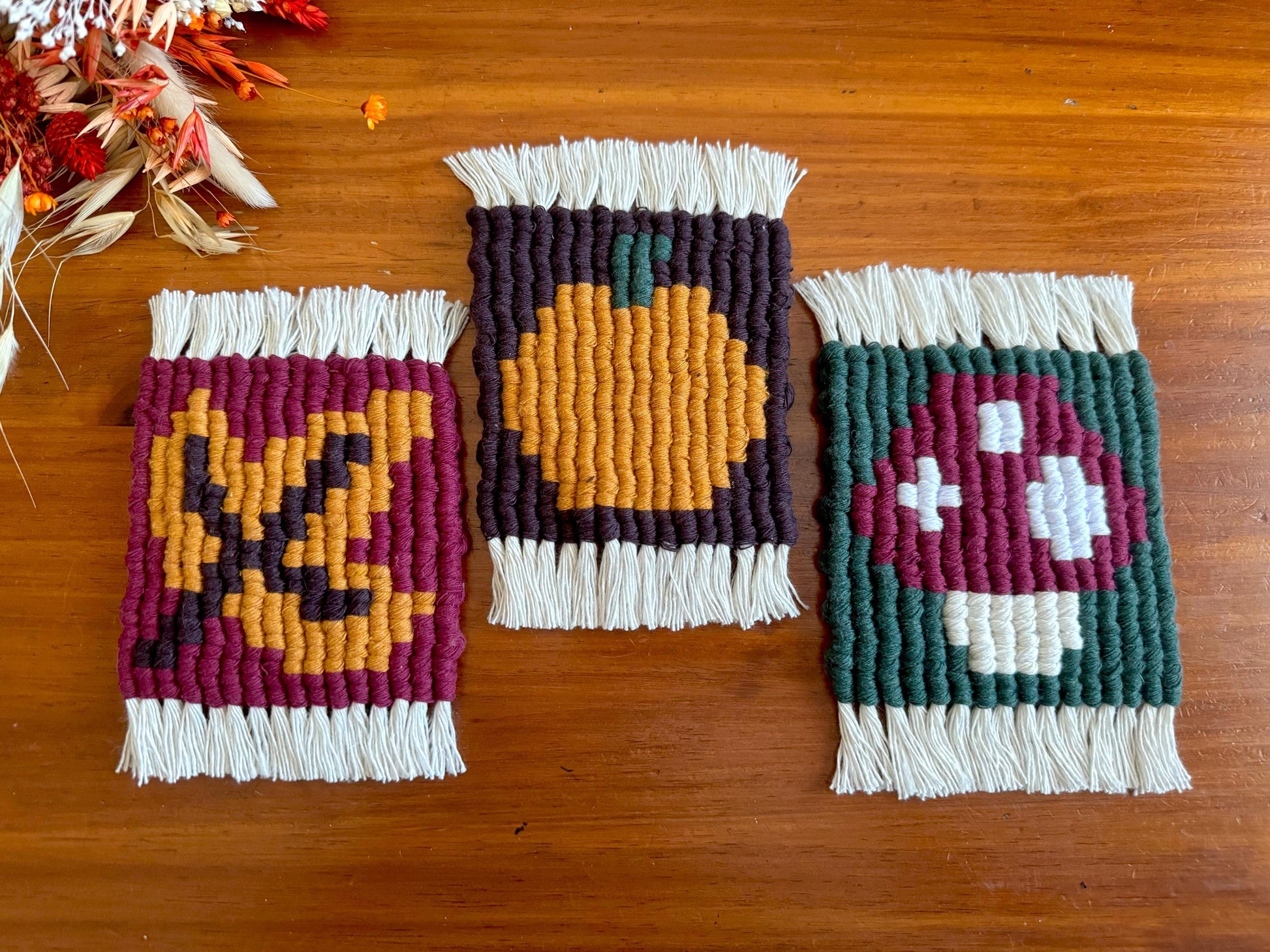 Three woven coasters with autumn-themed designs on a wooden surface.