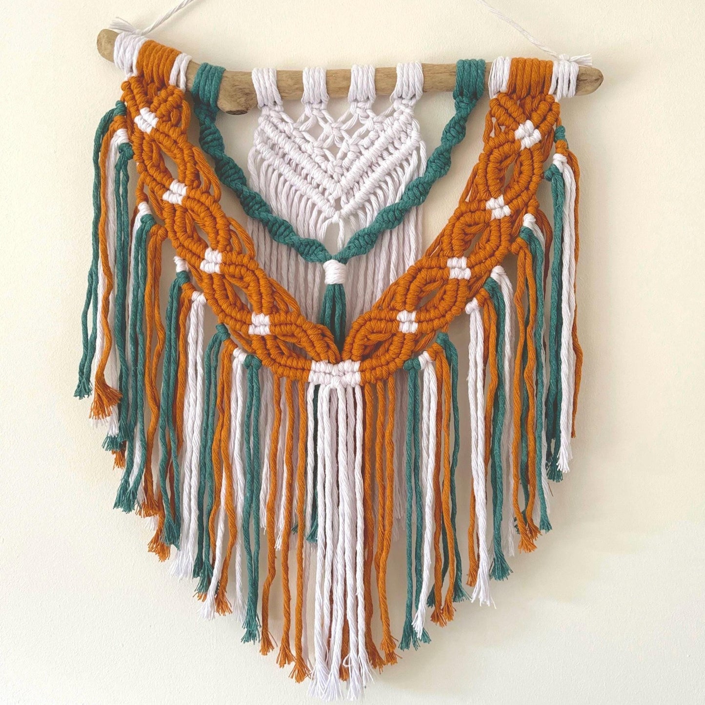 Macrame wall hanging with mustard, teal, and white ropes on a wooden stick against a white background.