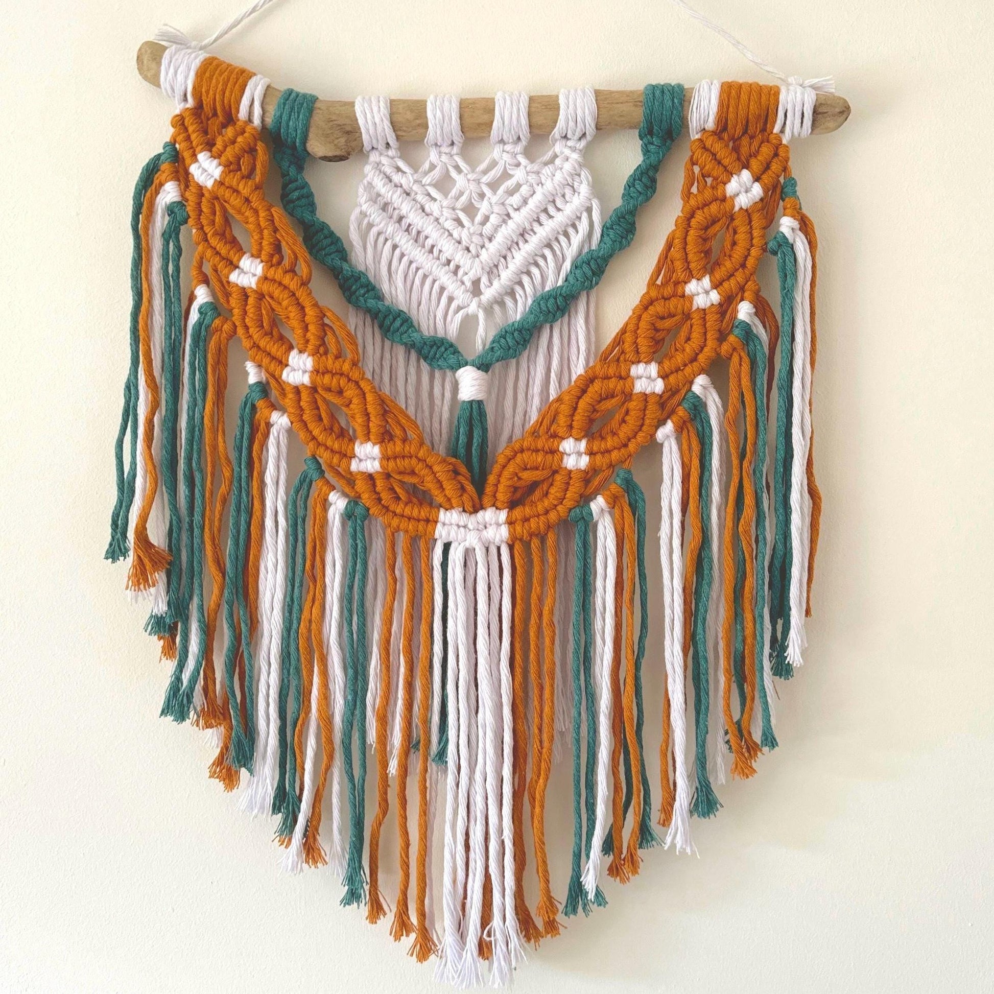 Macrame wall hanging with mustard, teal, and white ropes on a wooden stick against a white background.