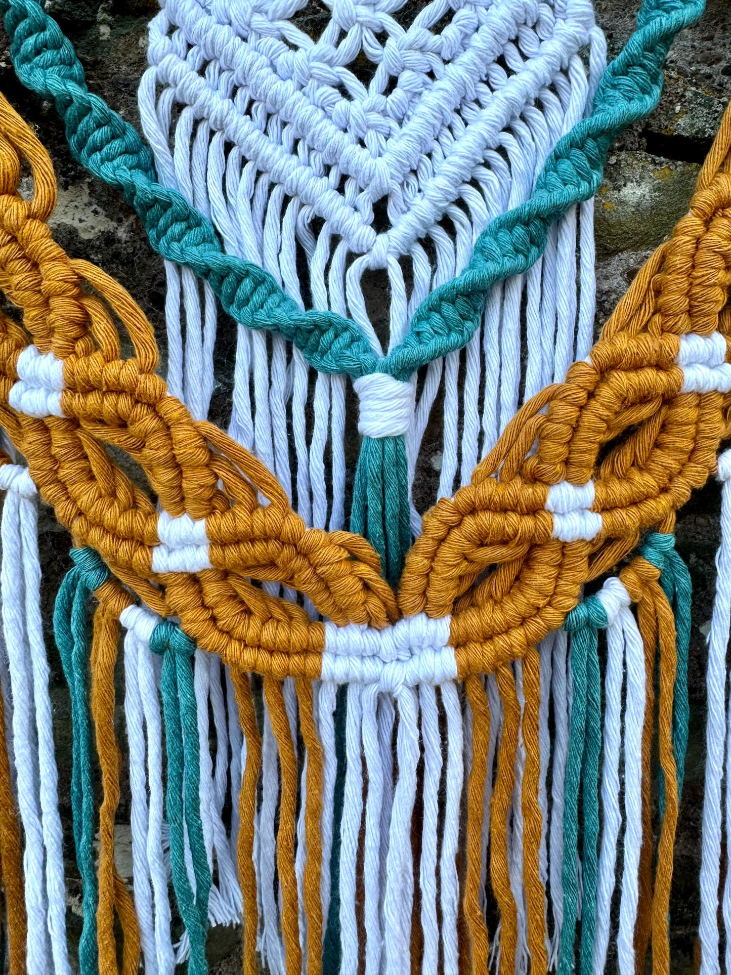 Close-up of a macrame wall hanging with mustard, teal, and white ropes.