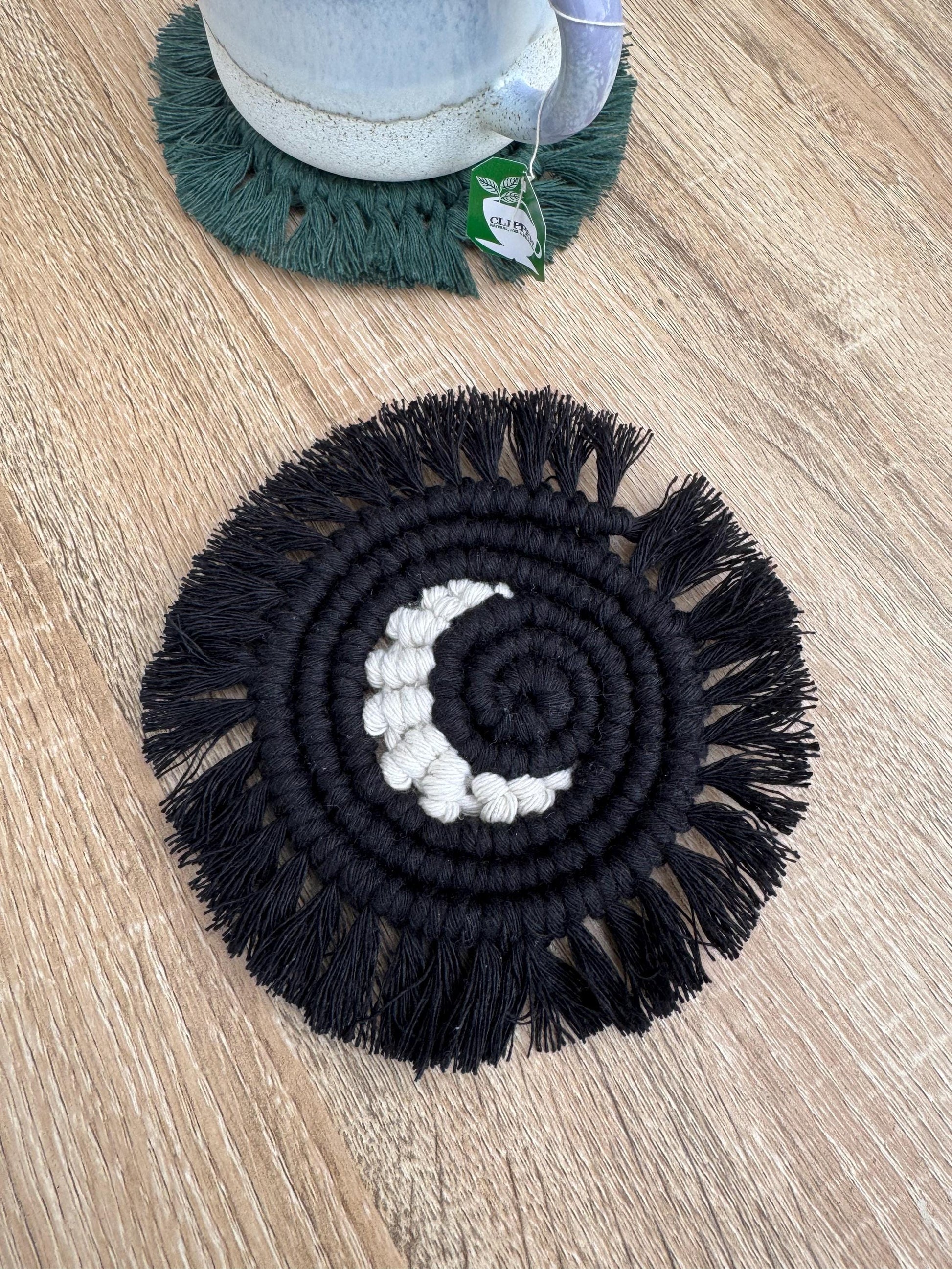 black macrame coaster with white moon detailing and a fringe on a wooden surface