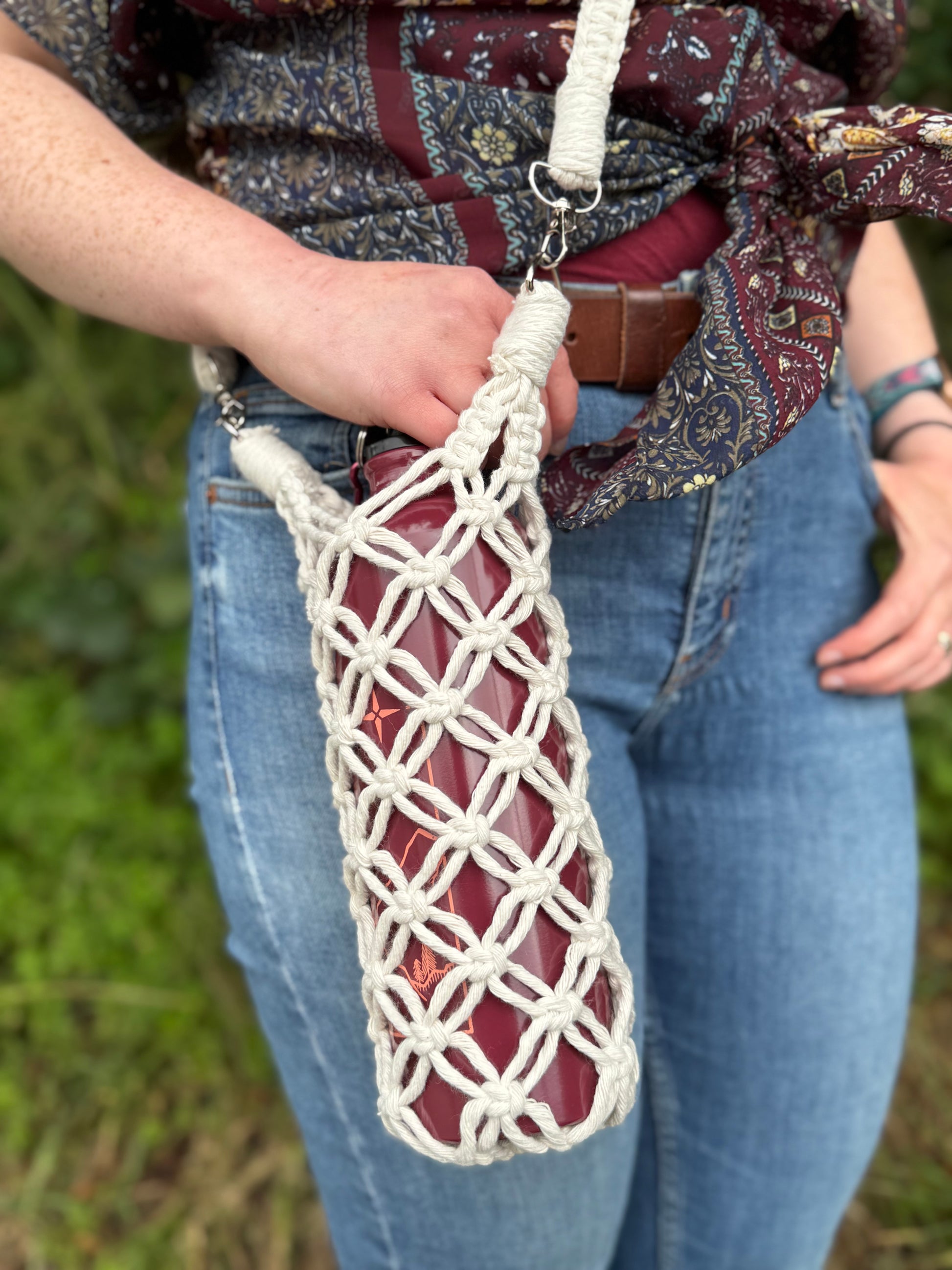 Person holding a macrame net bag with a red bottle against a blurred natural background