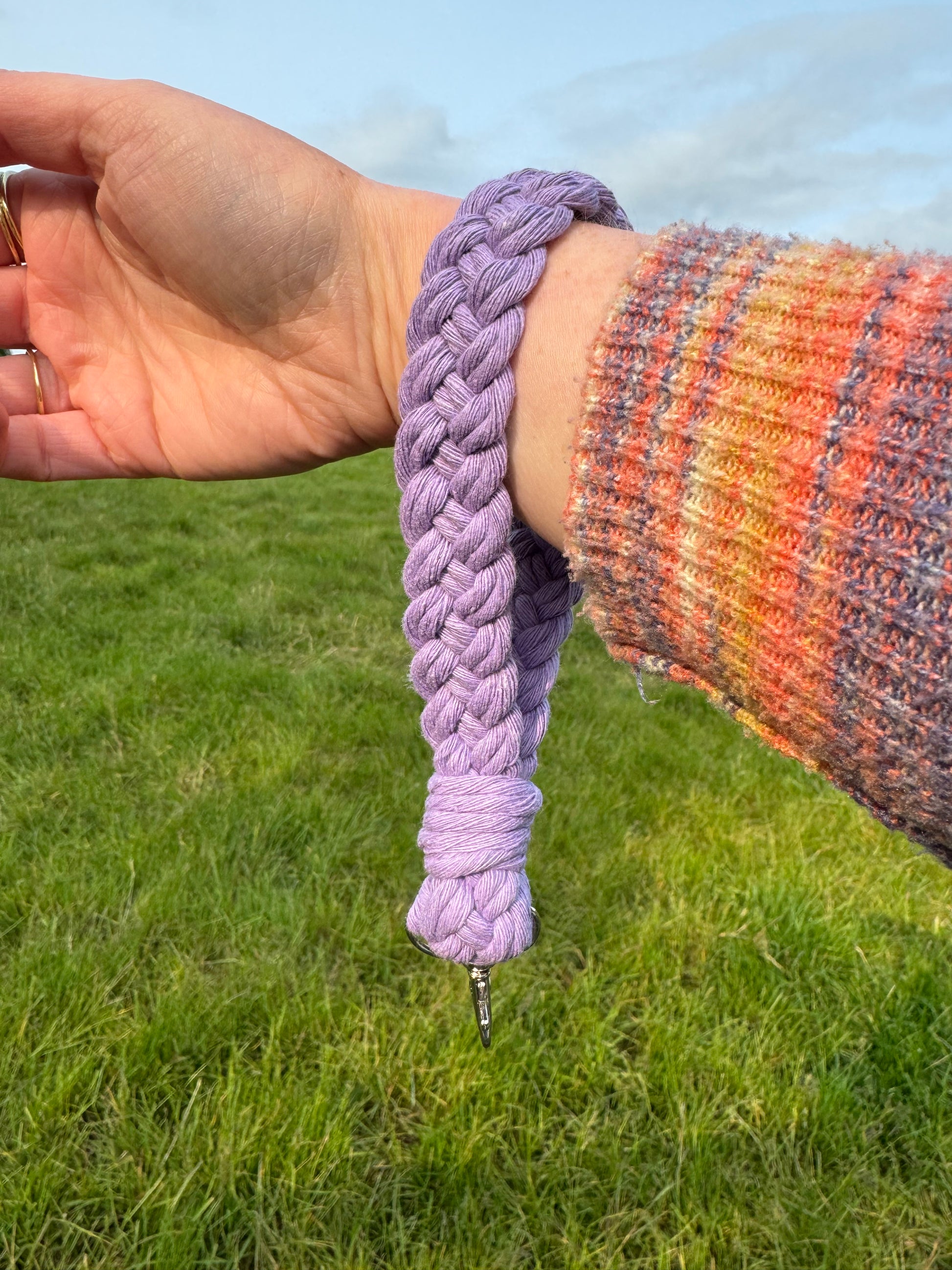 Hand wearing a braided purple keychain with a blurred background of grass and sky