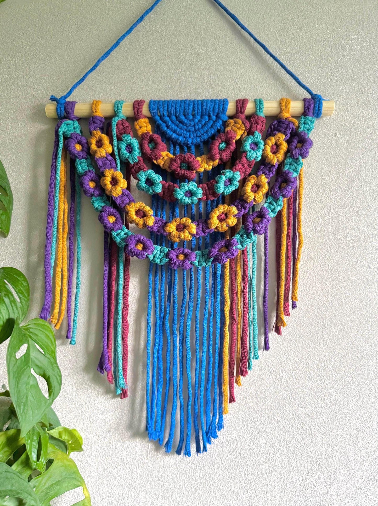 Colourful macrame wall hanging with floral patterns and long fringe on a white wall.