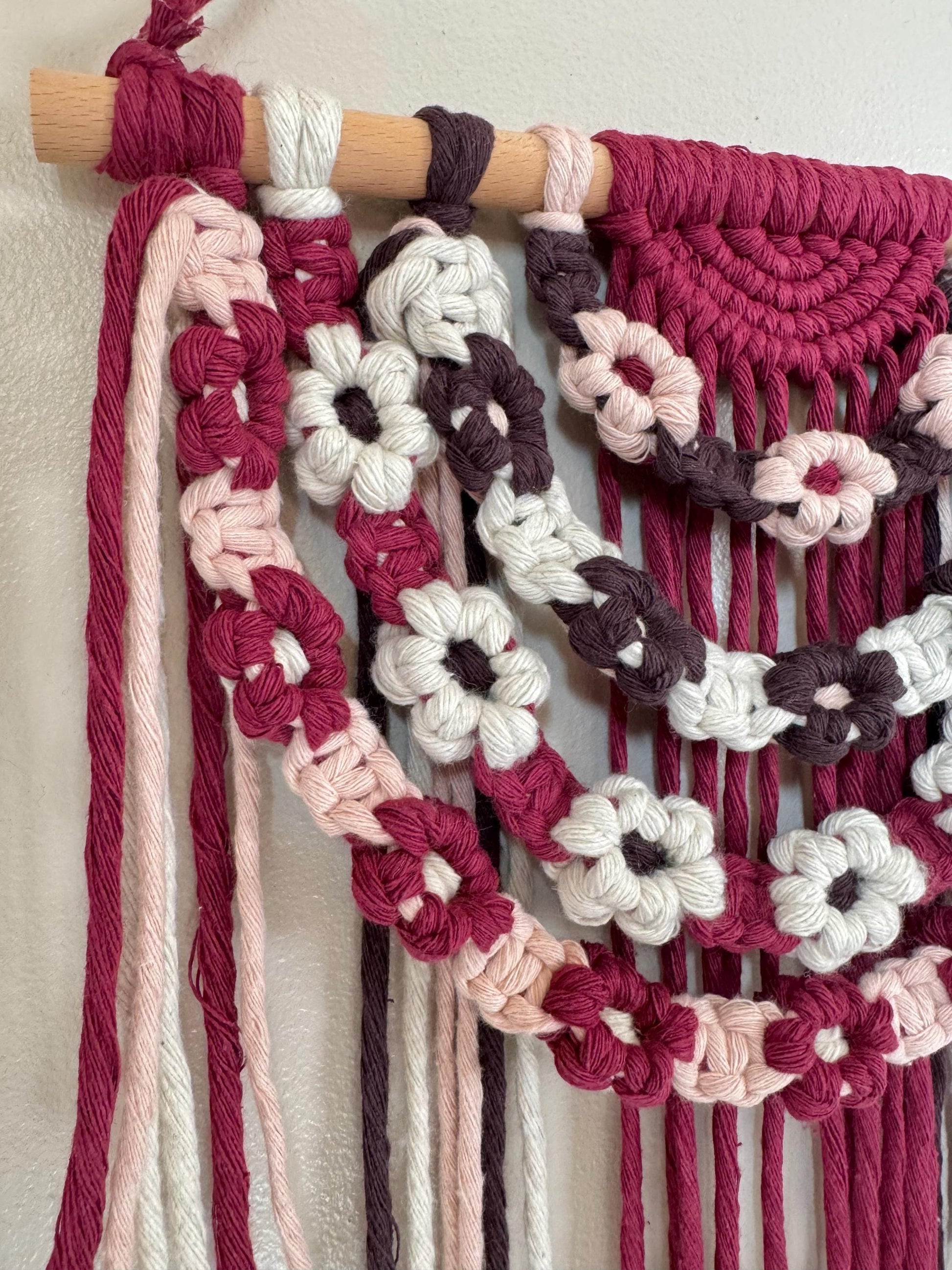 Macrame wall hanging with floral patterns in pink, white, and brown on a neutral background