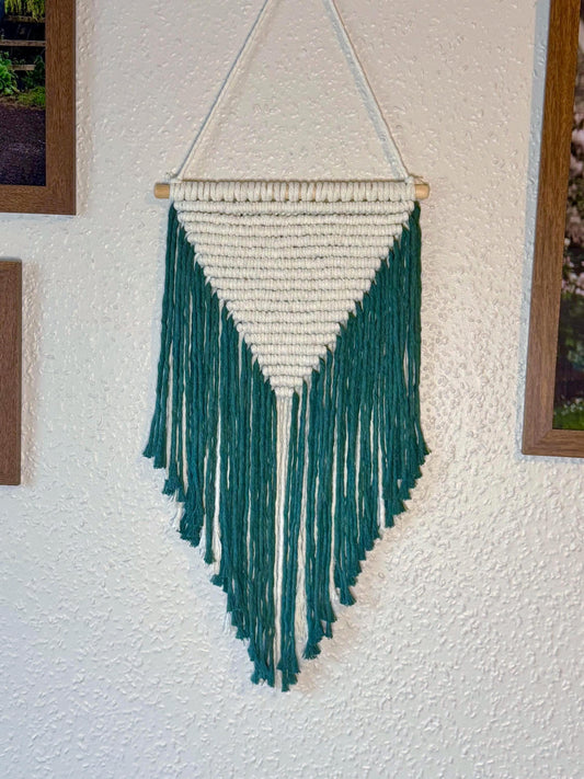 Macrame wall hanging with a natural-coloured triangular shape and a dark green fringe on a textured wall