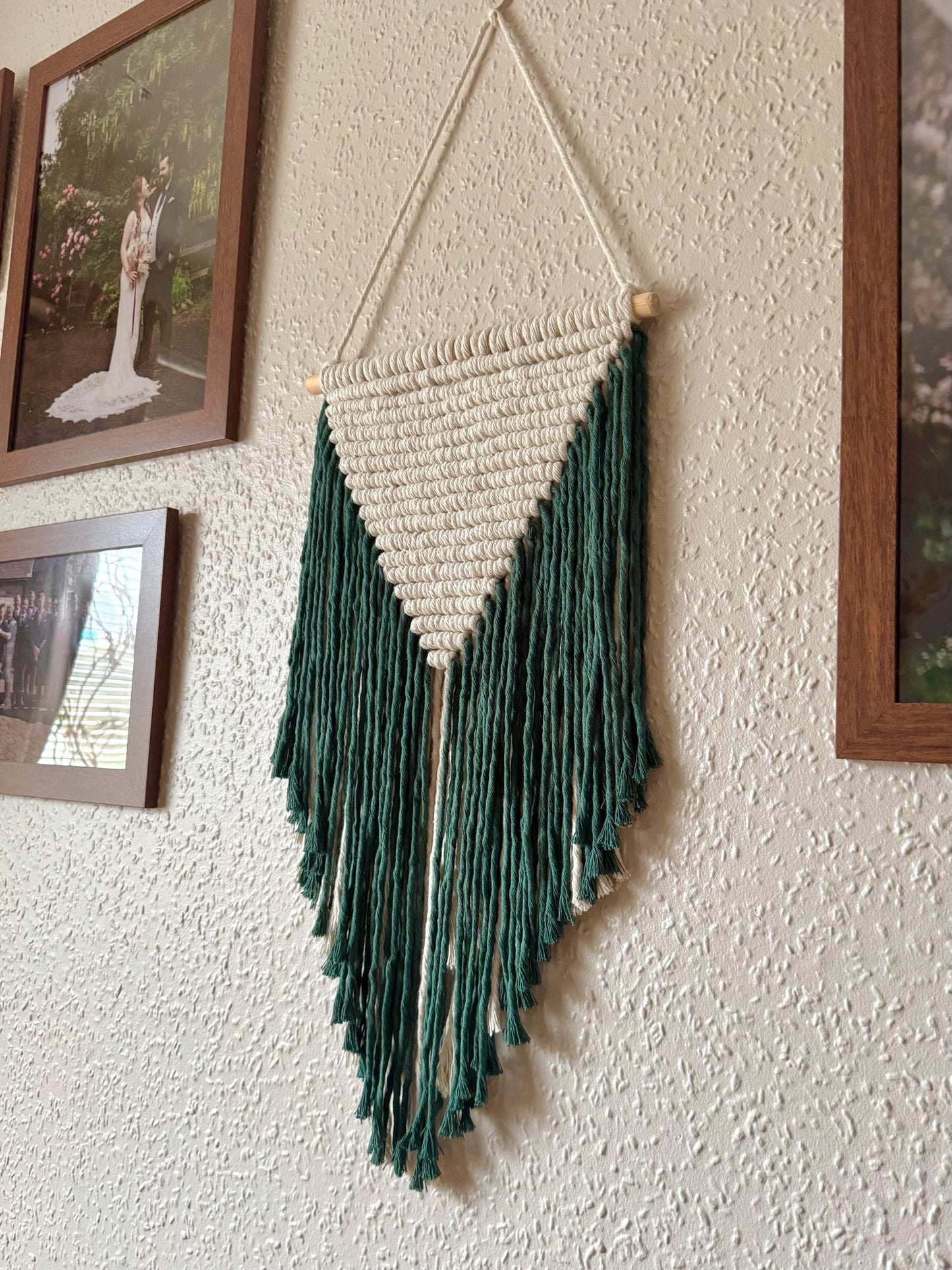 Macrame wall hanging with dark green fringe on a textured wall.