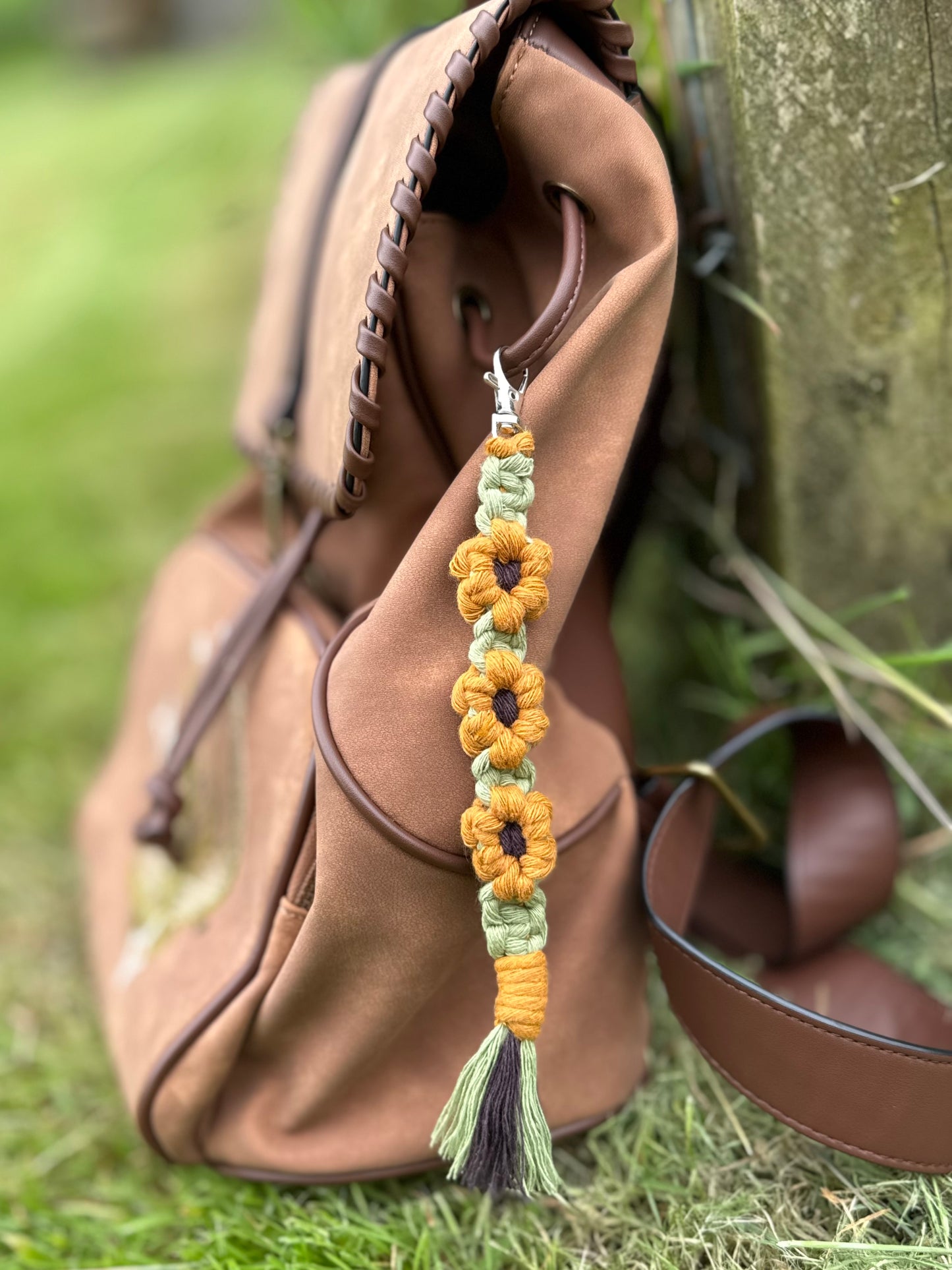 A decorative macrame sunflower keyring in colours yellow, brown and green, hanging from a brown backpack on the ground beside a treetrunk.