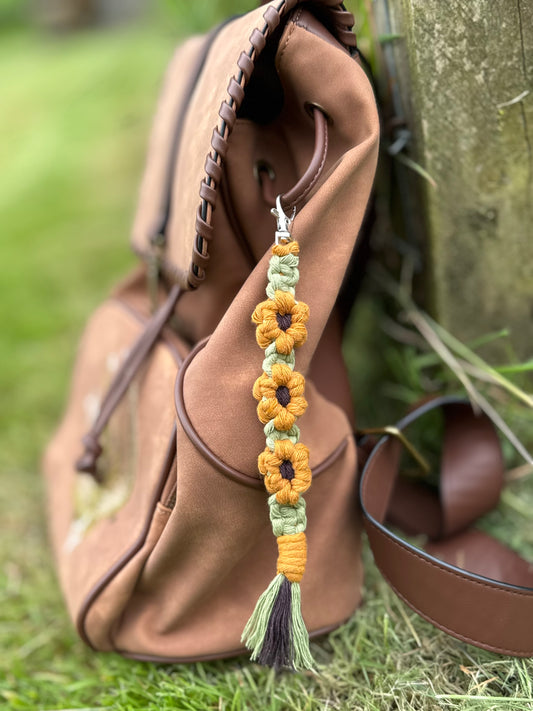 A decorative macrame sunflower keyring in colours yellow, brown and green, hanging from a brown backpack on the ground beside a treetrunk.