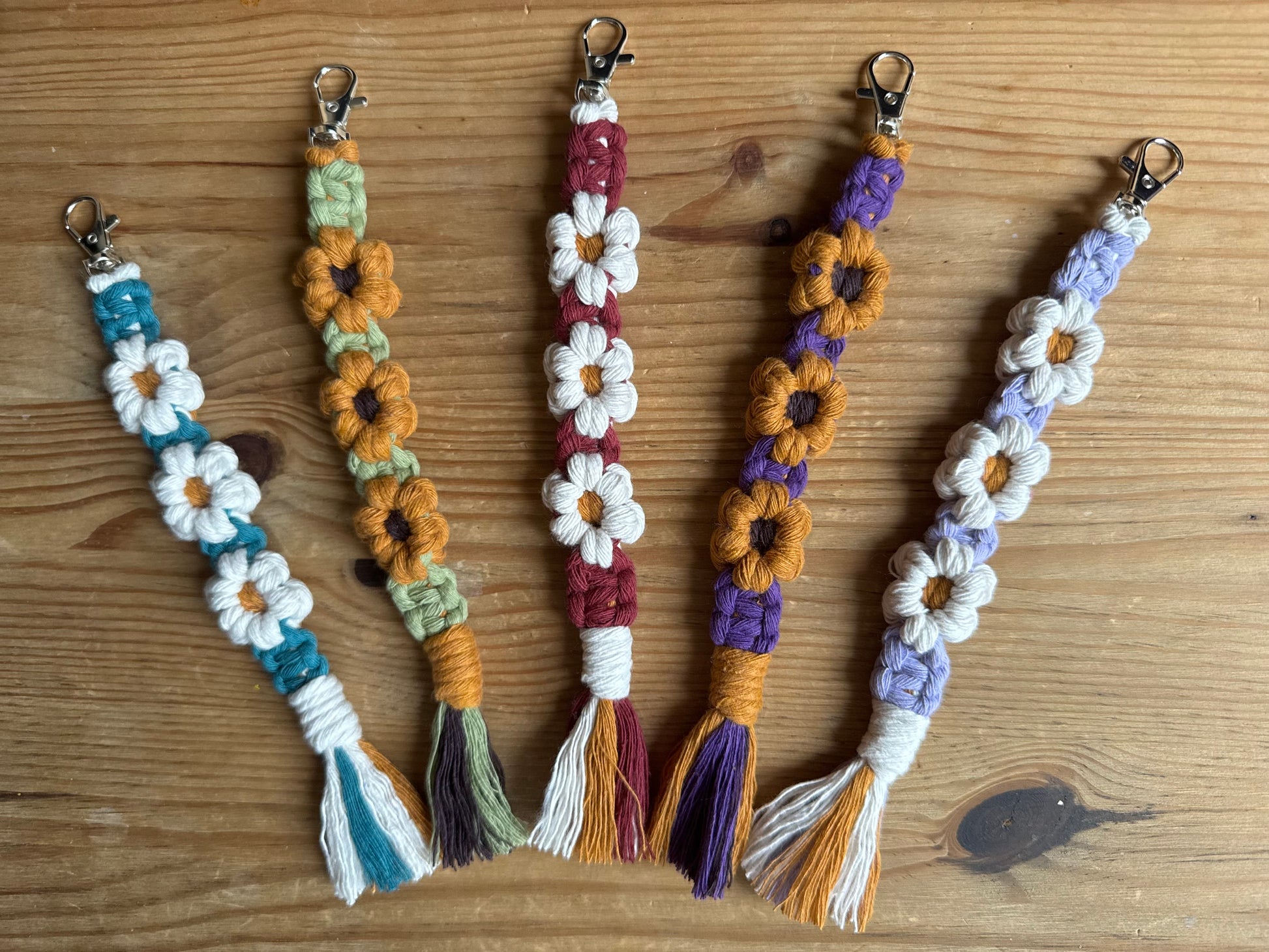 Five handmade macrame keychains with daisy and sunflower designs, arranged on a wooden surface. The keychains feature different color combinations including blue, green, orange, burgundy, purple, and lavender, each with a metal lobster clasp and tassel.