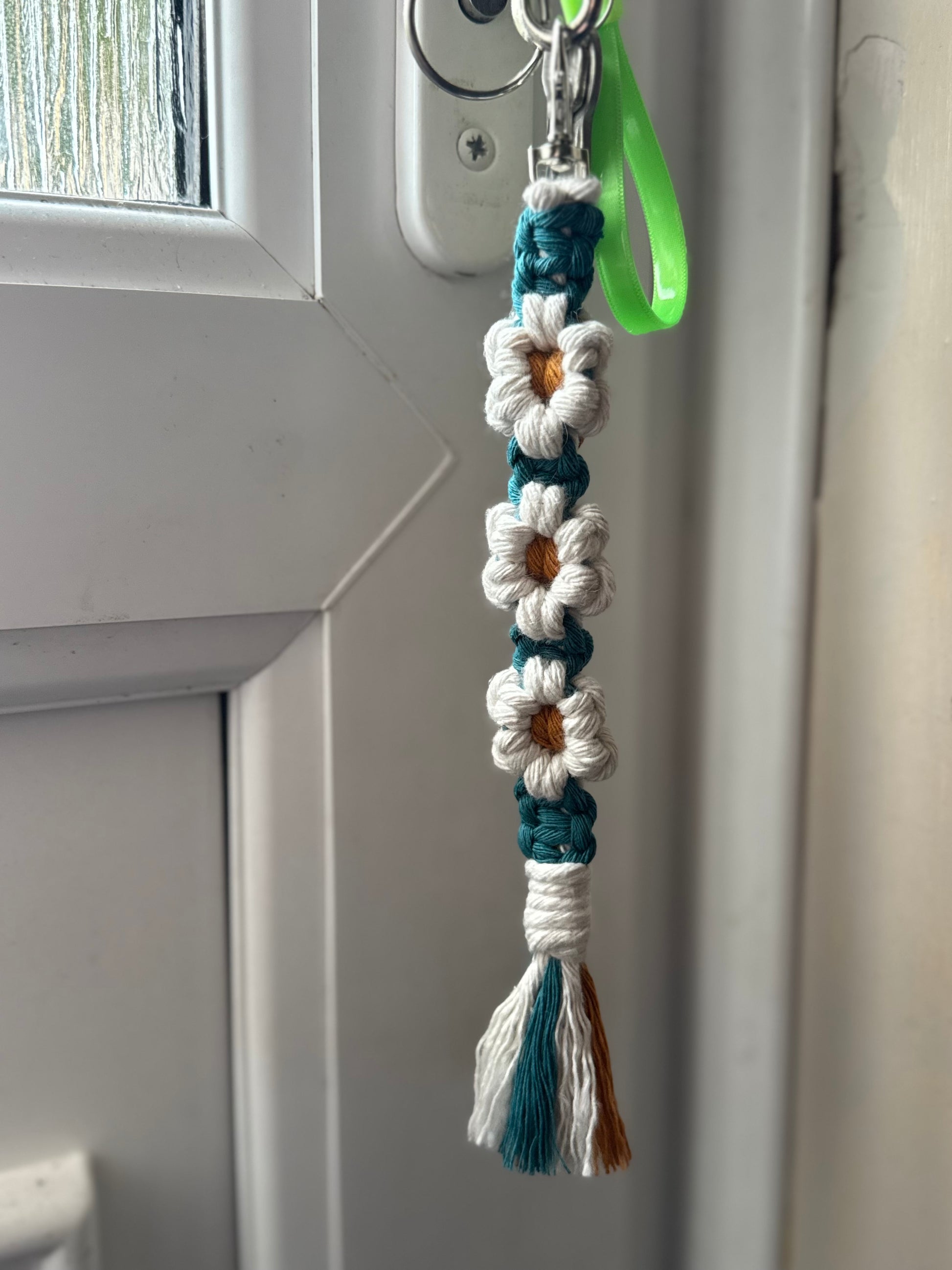 A handmade macrame keychain with white and teal woven flowers, a green wrist loop, and a tassel, hanging on a white door.