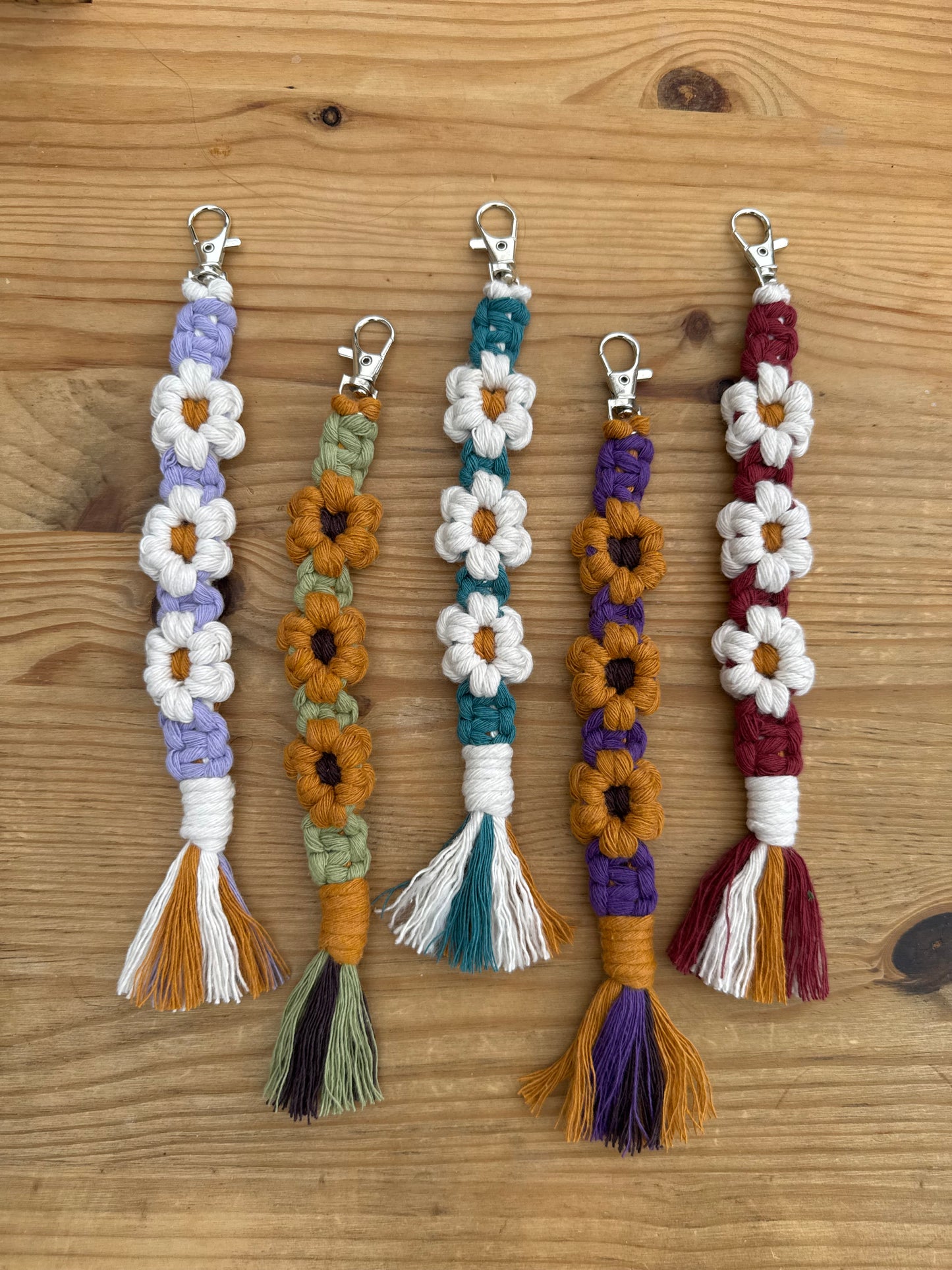 Five handmade macrame keychains with white daisy flowers and tassel ends, laid out horizontally on a light brown wooden surface. Each keychain features a different color combination: burgundy, purple, teal, and lavender, paired with white, mustard, and olive accents.