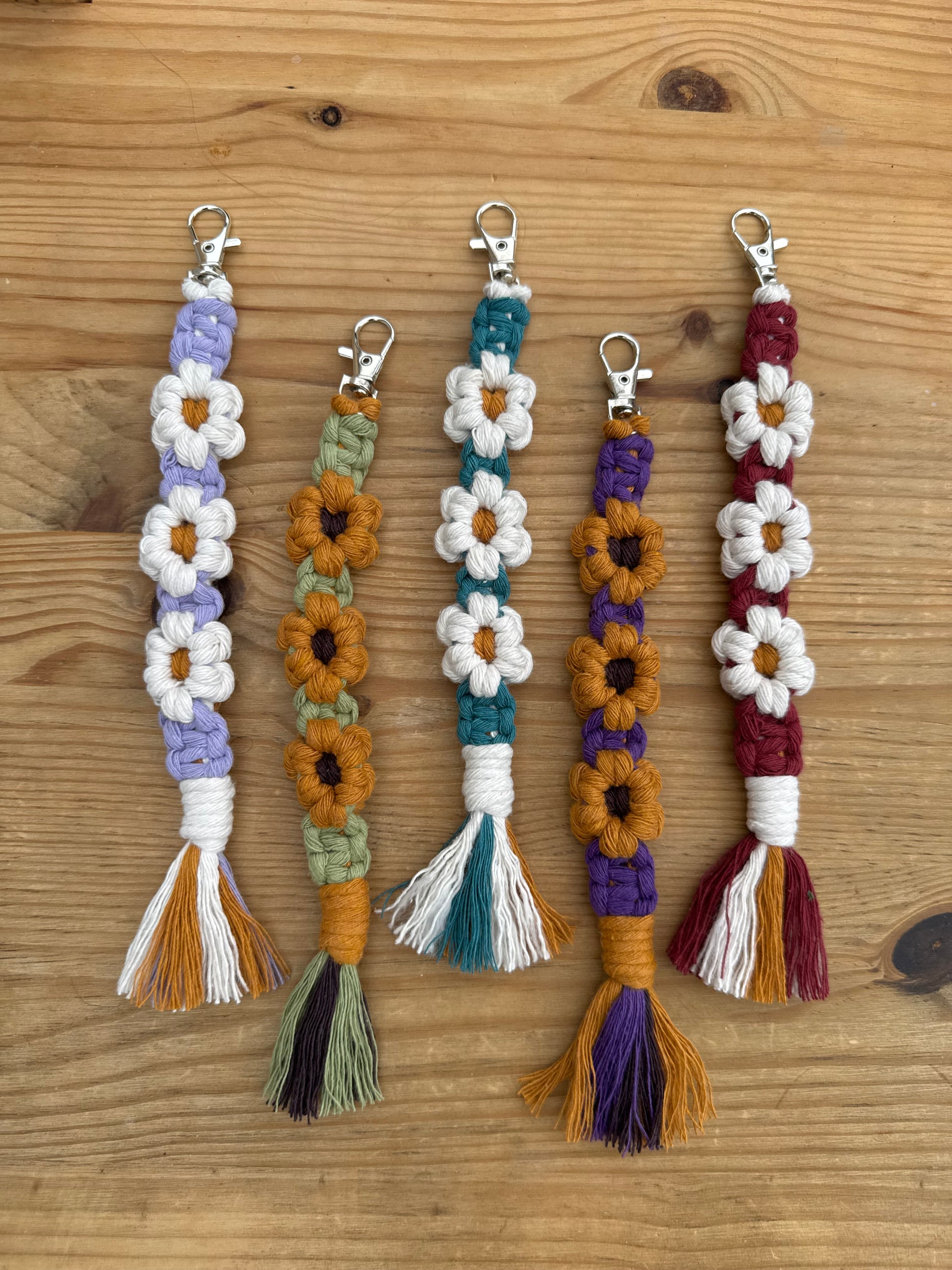Five handmade macrame keychains with white daisy flowers and tassel ends, laid out horizontally on a light brown wooden surface. Each keychain features a different color combination: burgundy, purple, teal, and lavender, paired with white, mustard, and olive accents.