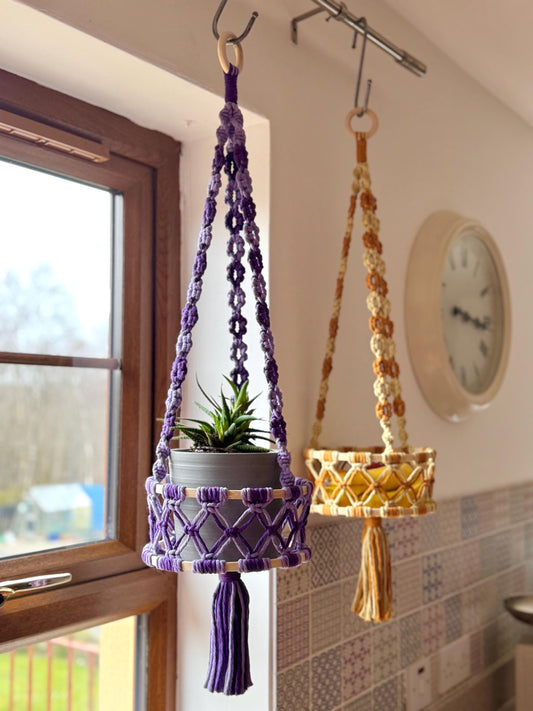 Two macrame plant hangers, one purple and one yellow, hanging from hooks on a wall.