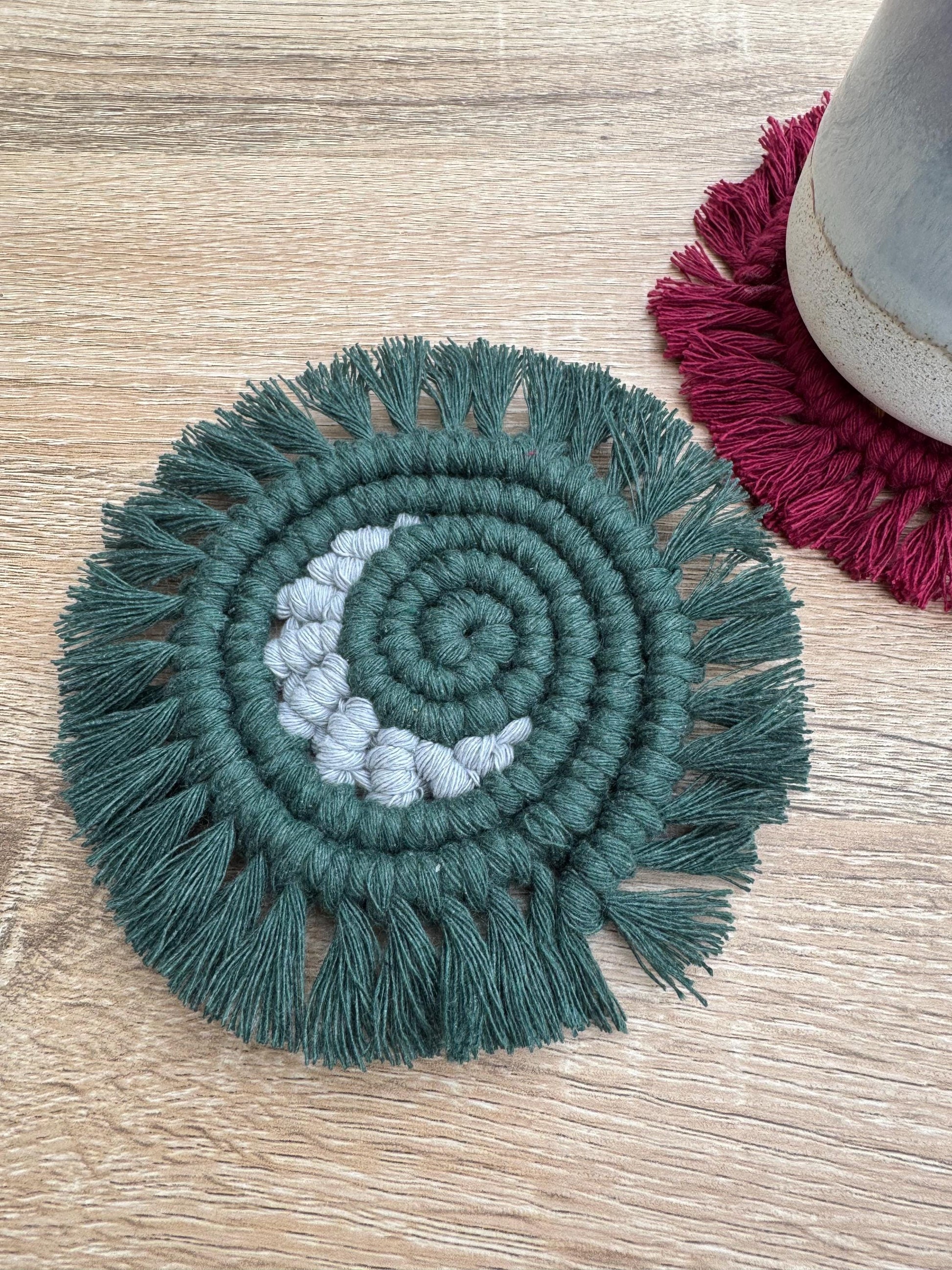 Green circular macrame coaster with a grey crescent moon and with fringe on a wooden surface