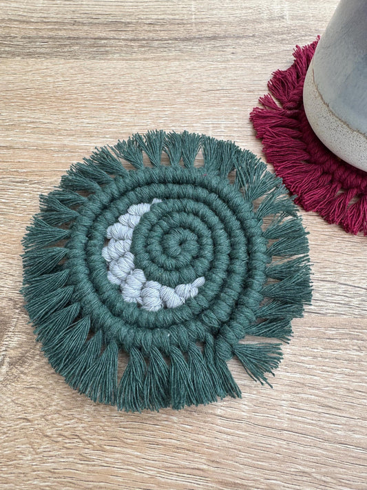 Green circular macrame coaster with a grey crescent moon and with fringe on a wooden surface