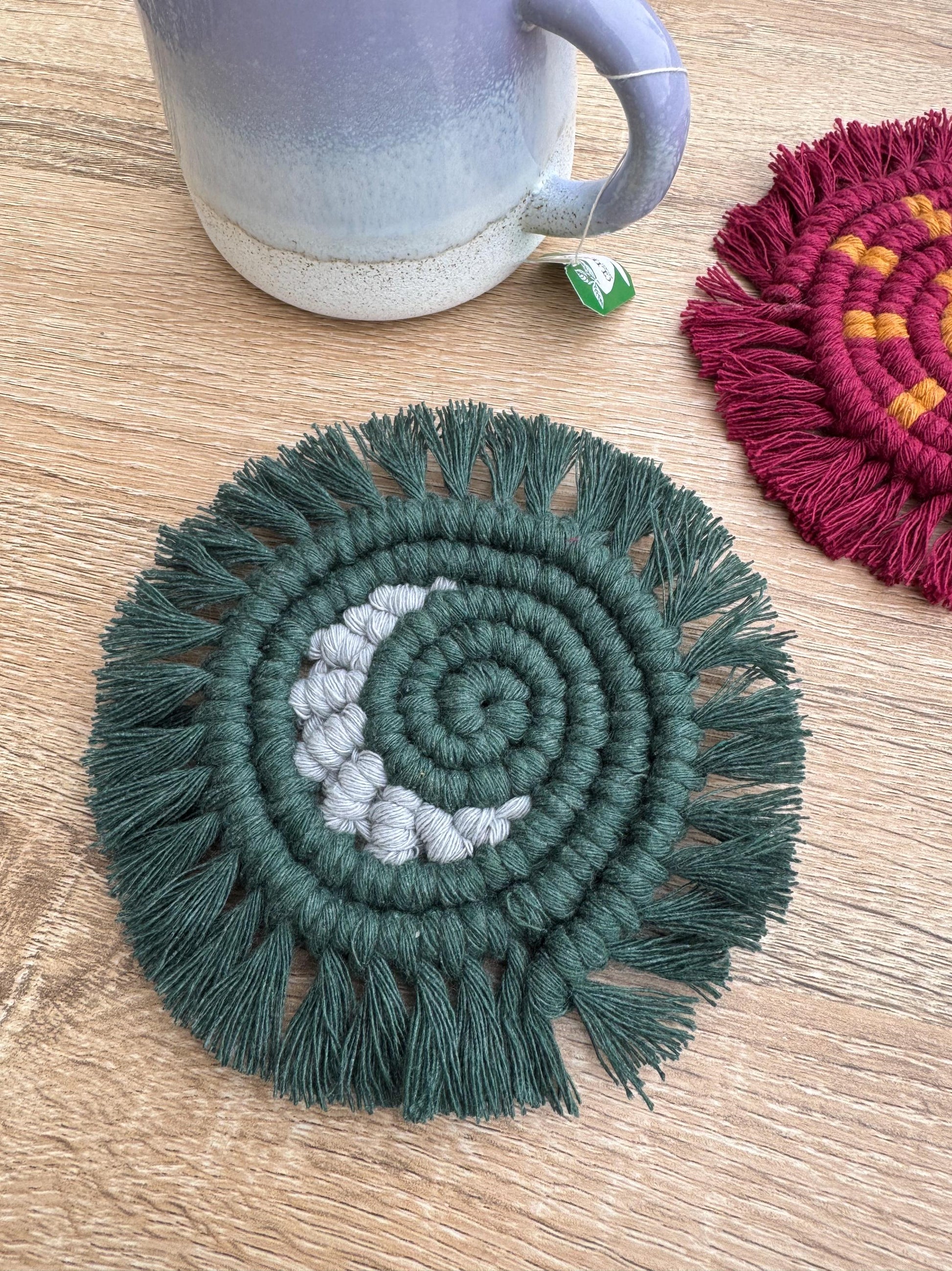 Green and grey macrame moon coaster on a wooden surface with a mug and another coaster in the background.