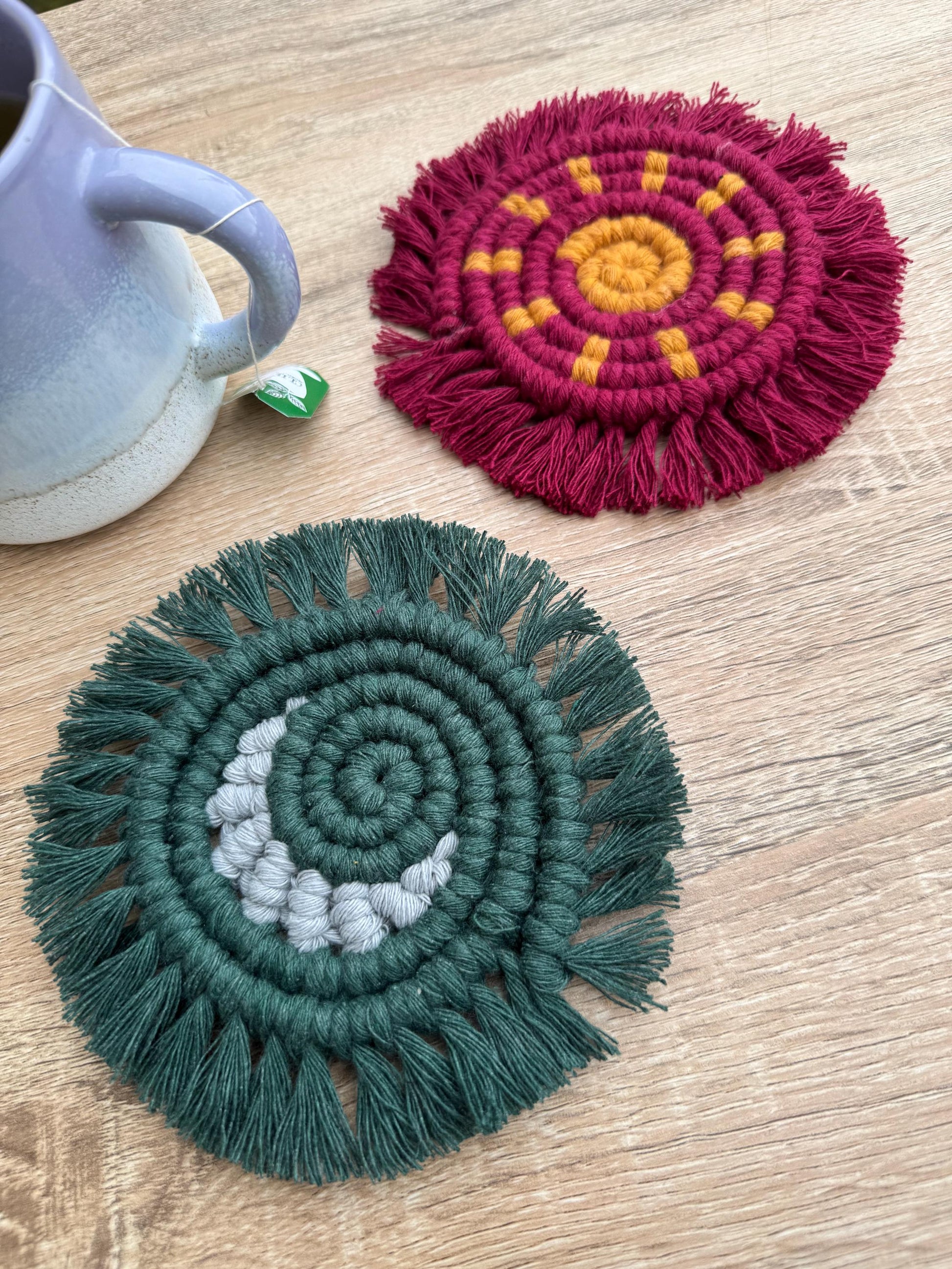 Two macrame coasters, one green with grey accents and one red with yellow accents, on a wooden surface.