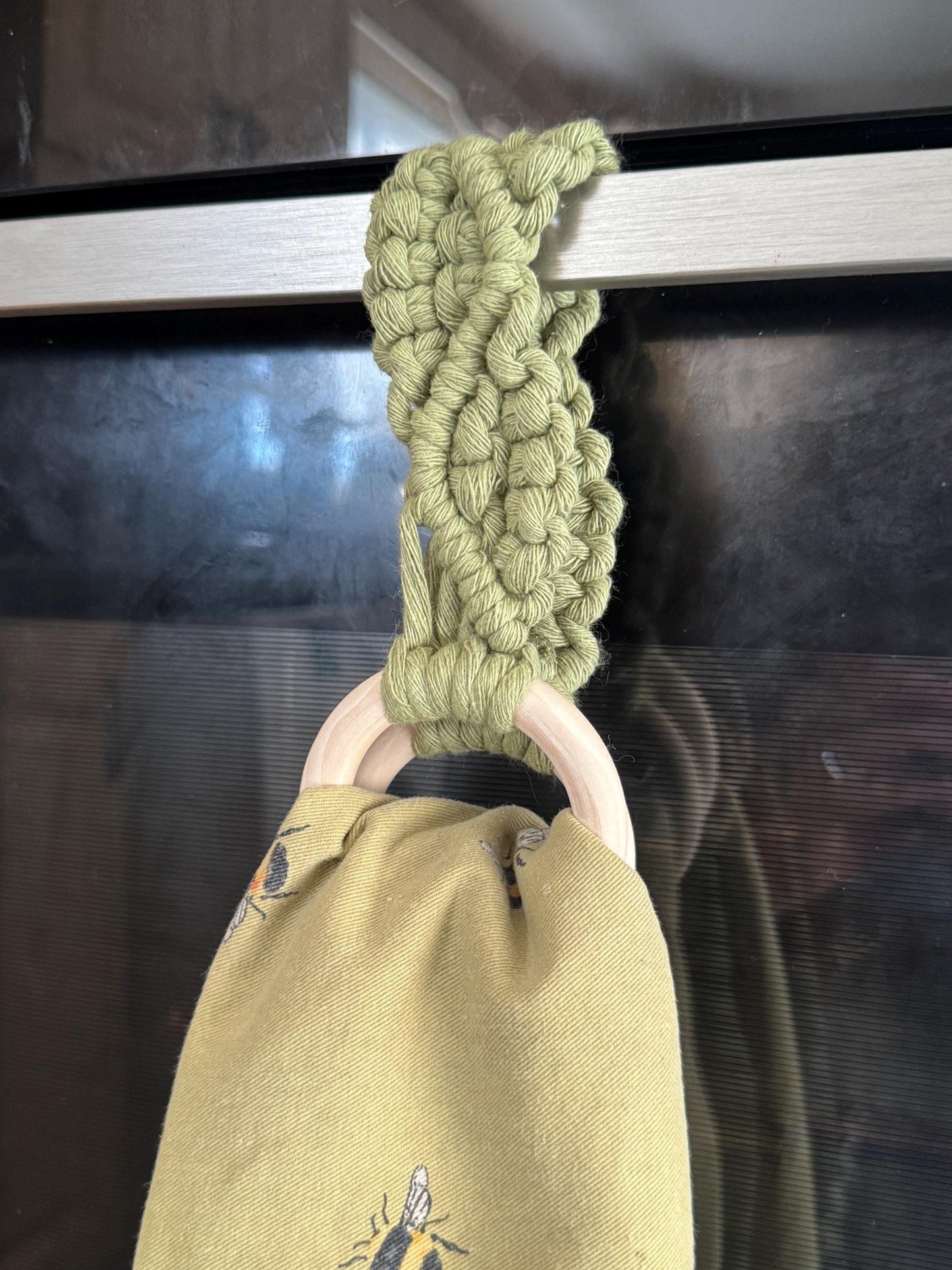 Green macrame kitchen towel holder with a wooden ring, holding a yellow tea towel on an oven door