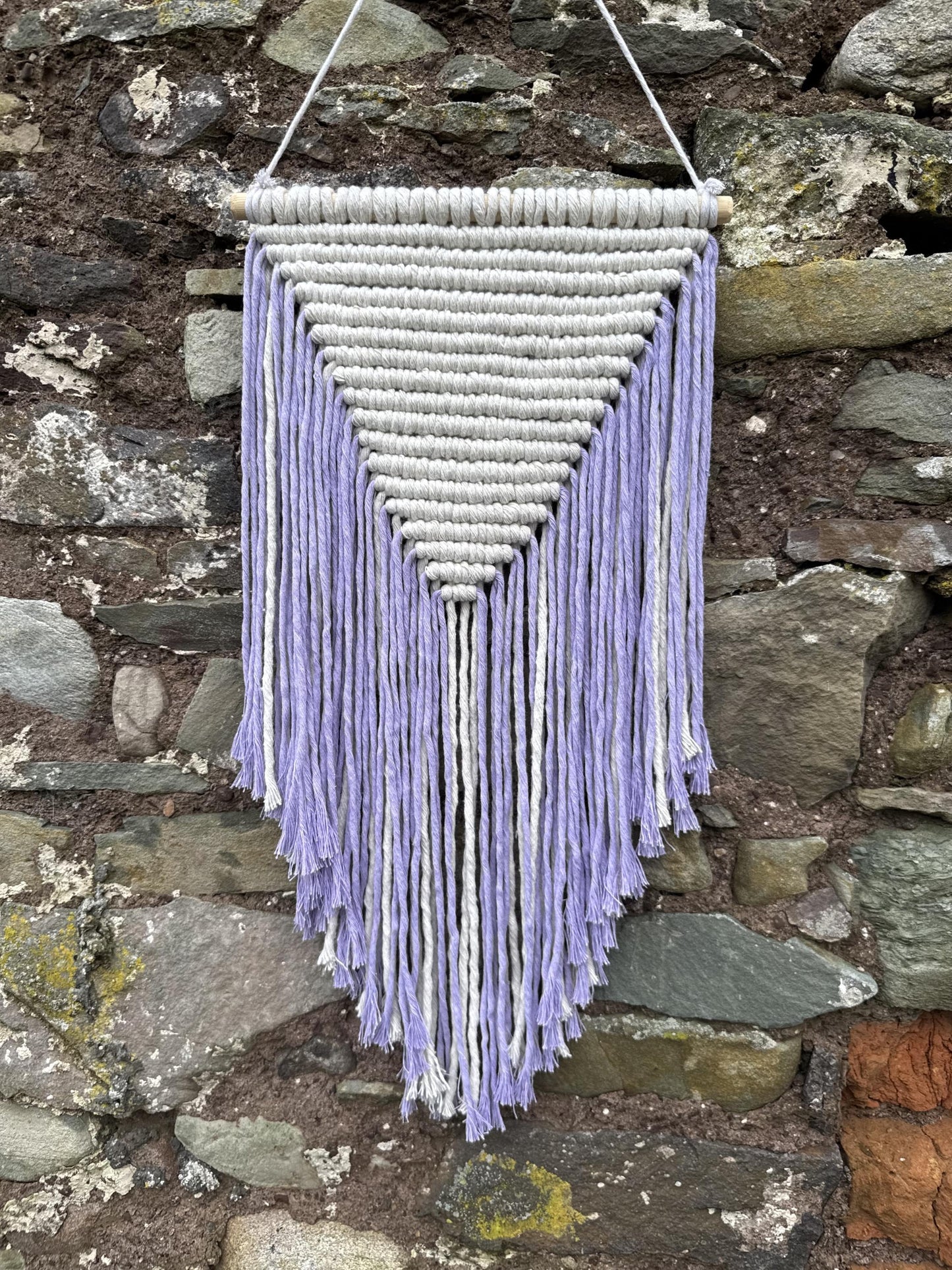 Macrame wall hanging with purple and white strings against a stone wall.