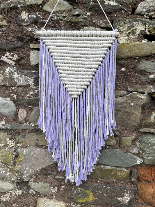 Macrame wall hanging with purple and white strings against a stone wall.
