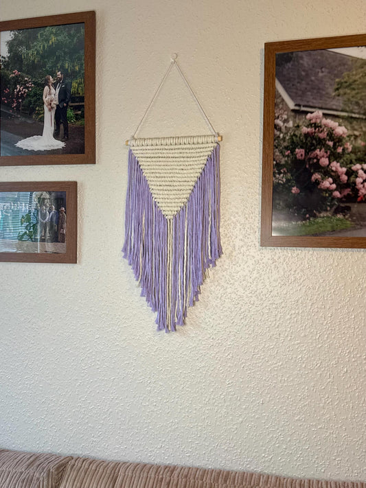 Decorative wall hanging with purple fringe on a textured wall with framed pictures.