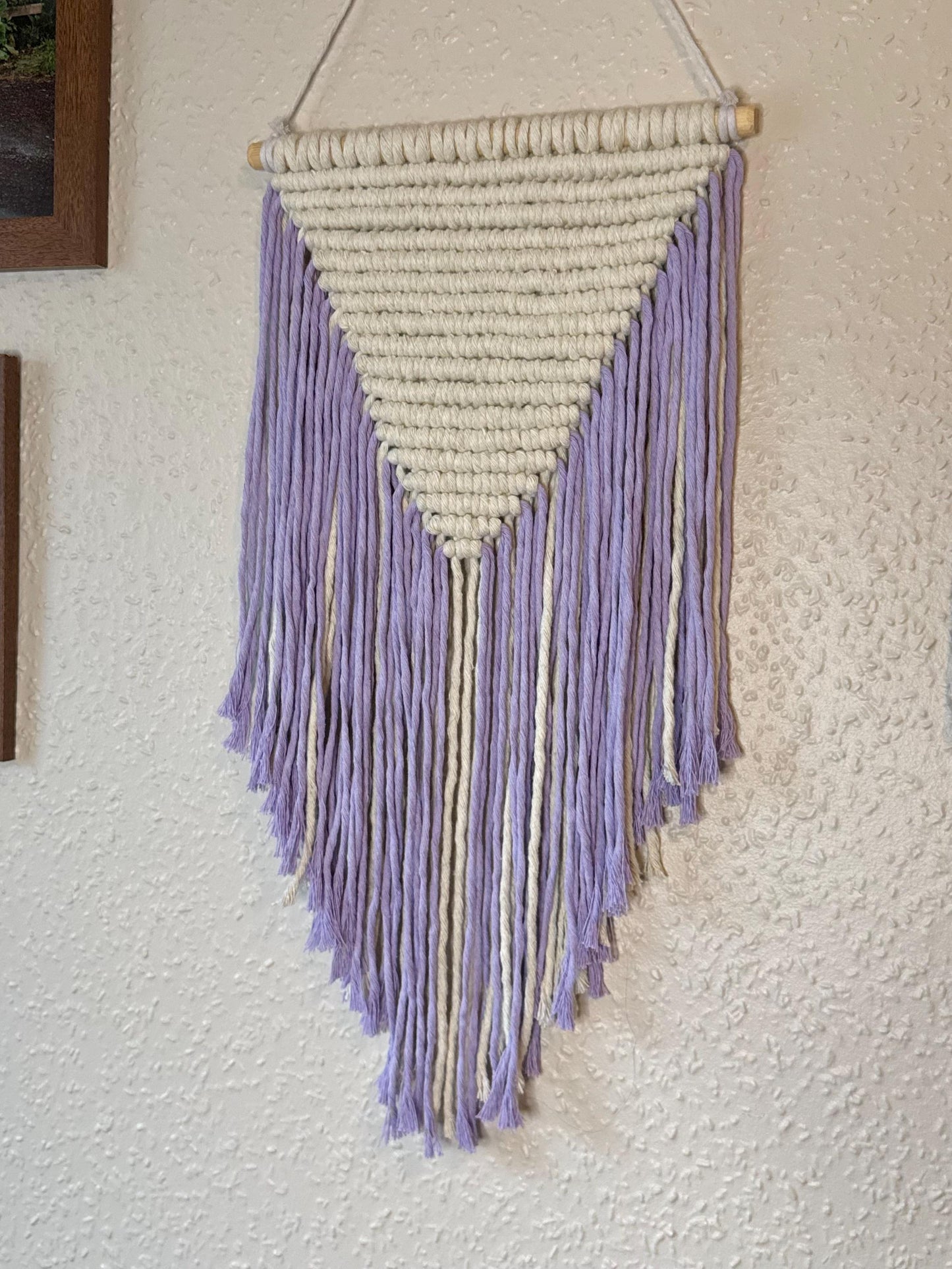 Macrame geometric wall hanging with lavender and white fringes on a textured wall.