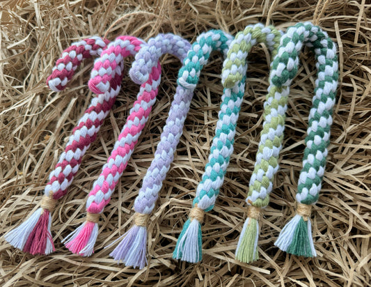 six macrame candy cane ornaments in different colours on a bed of shredded brown paper