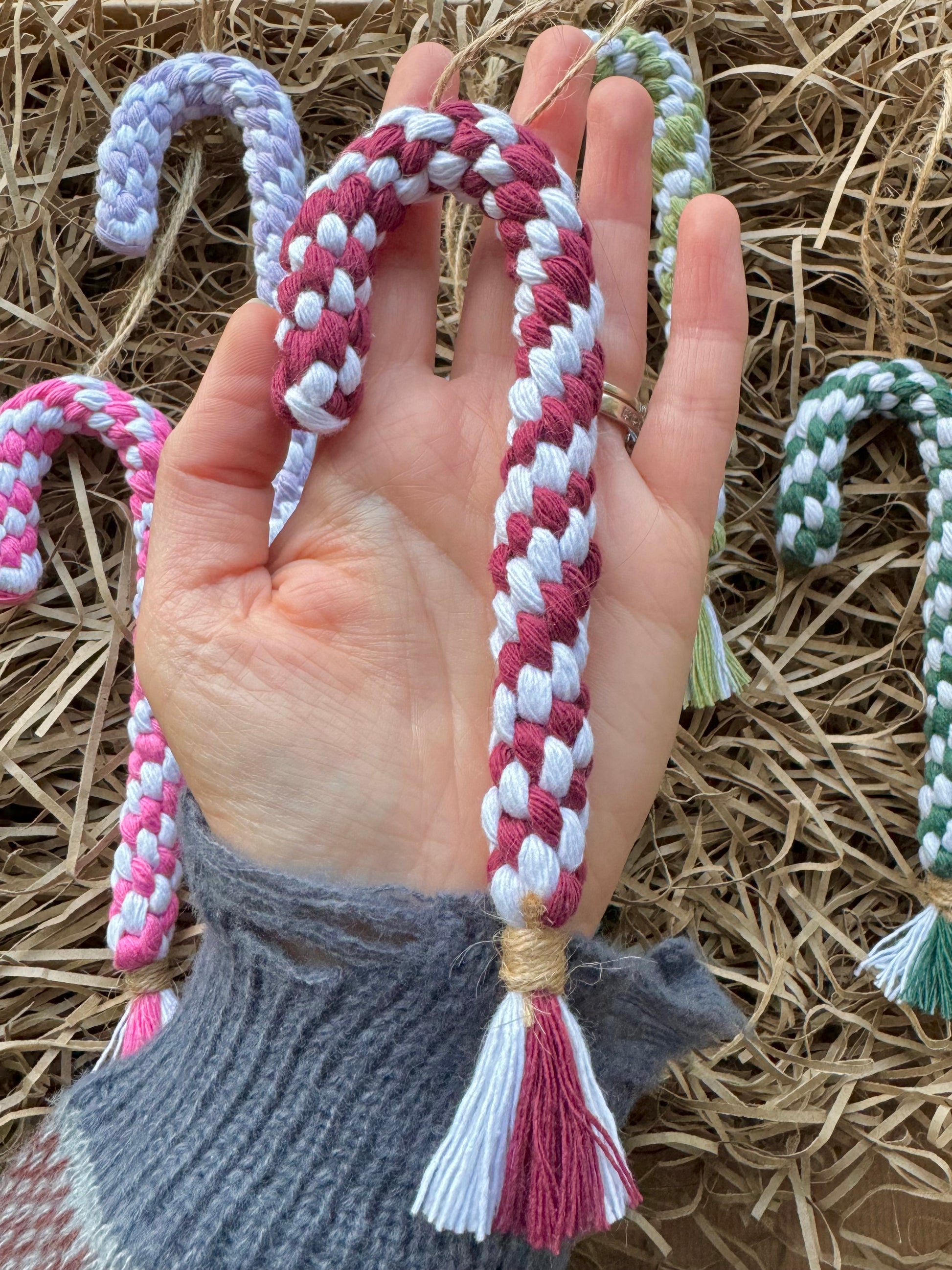 Hand holding a braided candy cane decoration with colorful tassels on a straw background