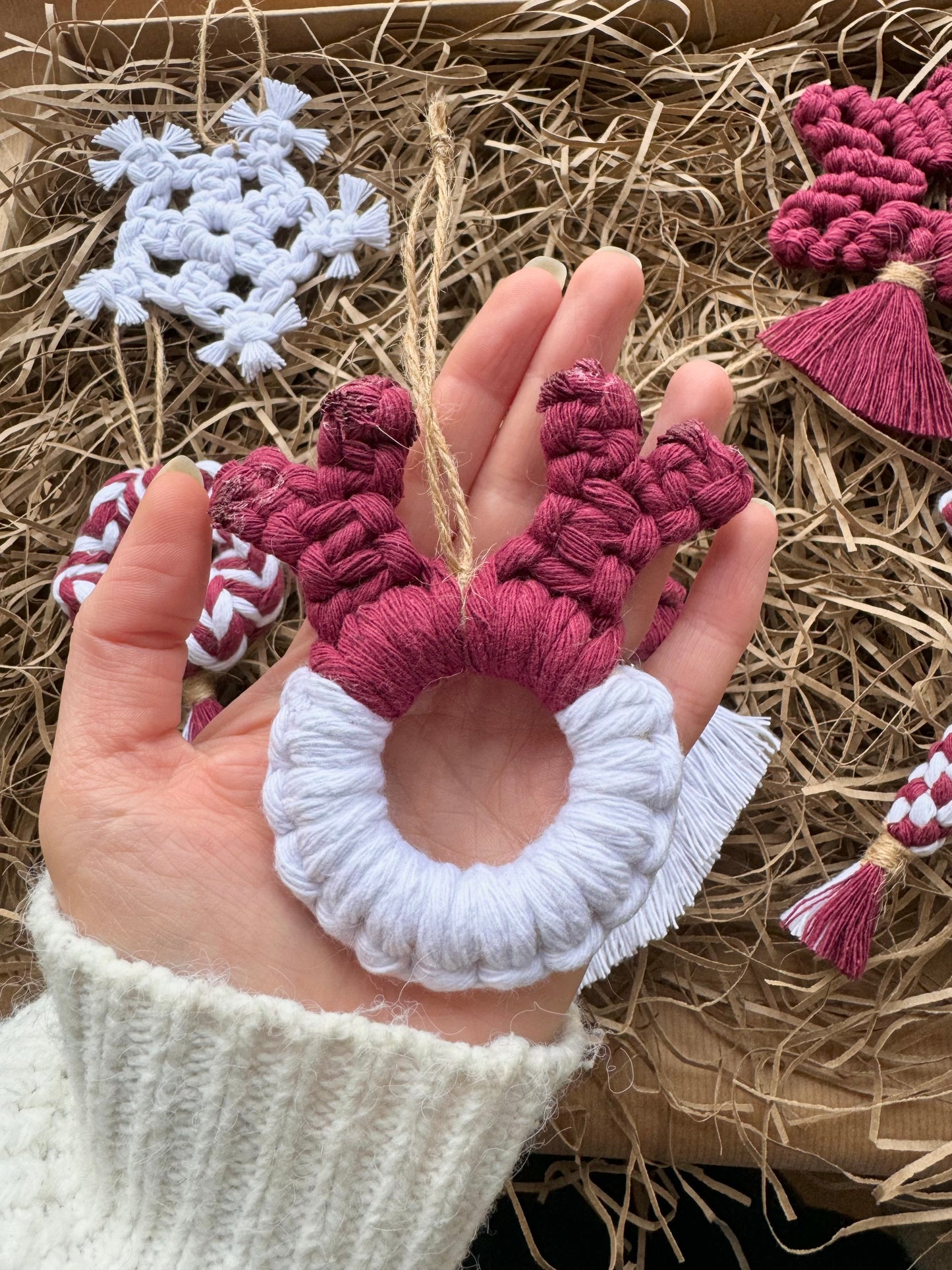 Hand holding a white and burgundy macrame reindeer ornament with decorative items in the background.