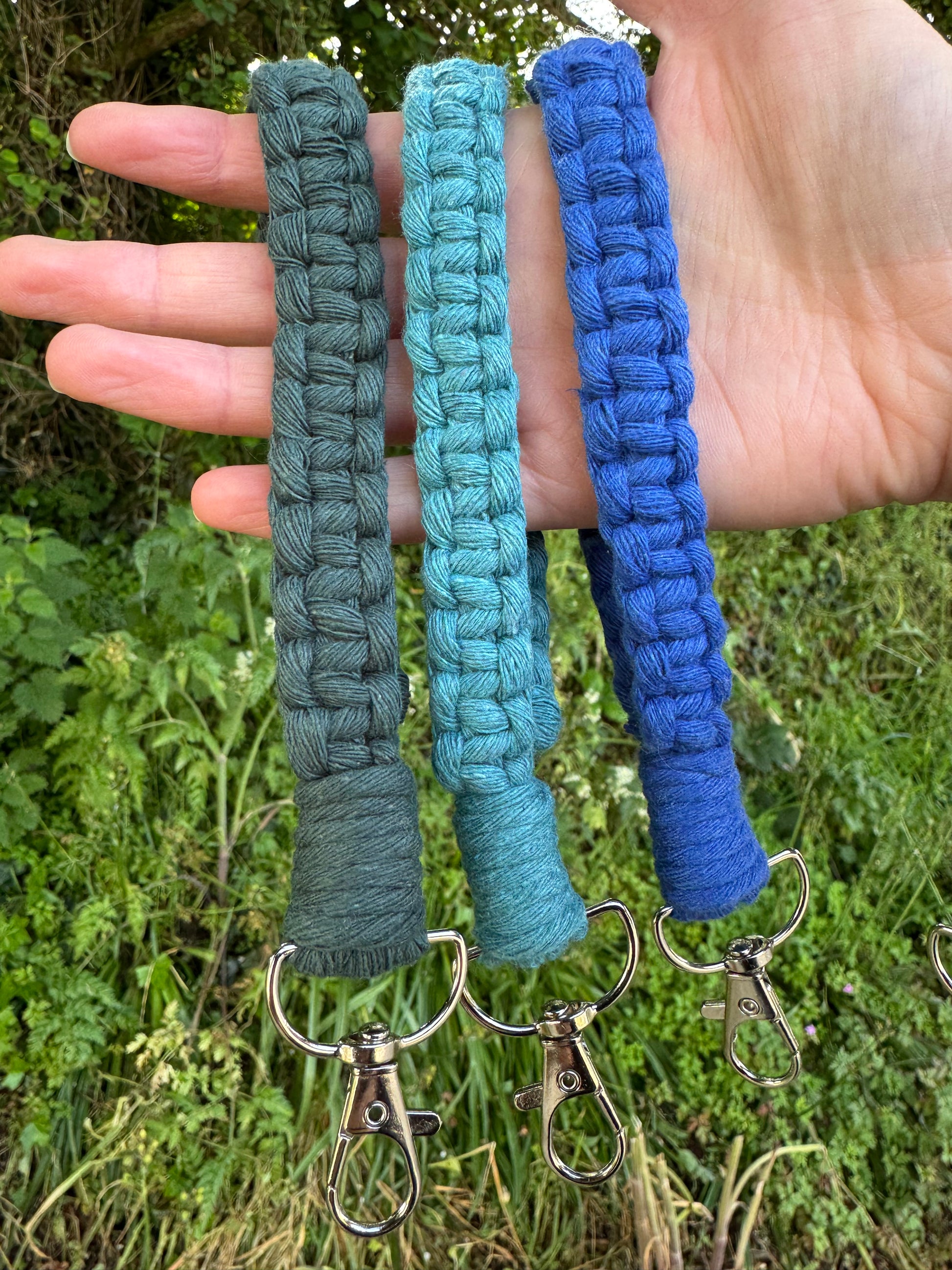 Three braided keychains in blue, green, and gray held against a grassy background.