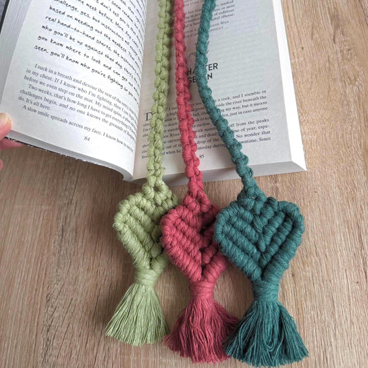 Three macrame heart bookmarks in light green, terracotta, and dark green on an open book.