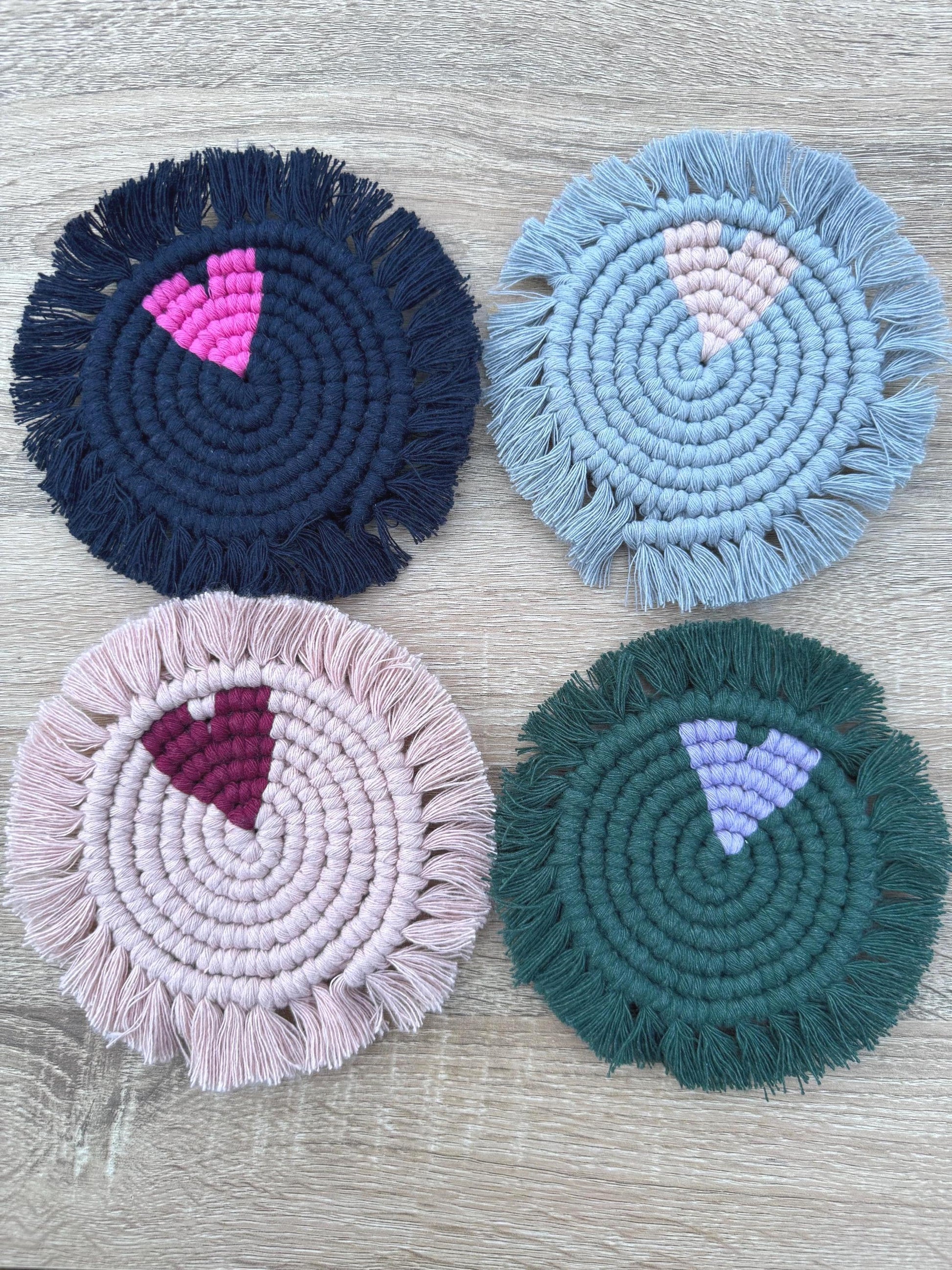 Four round macrame coasters with heart features and fringes in different colors on a wooden surface.