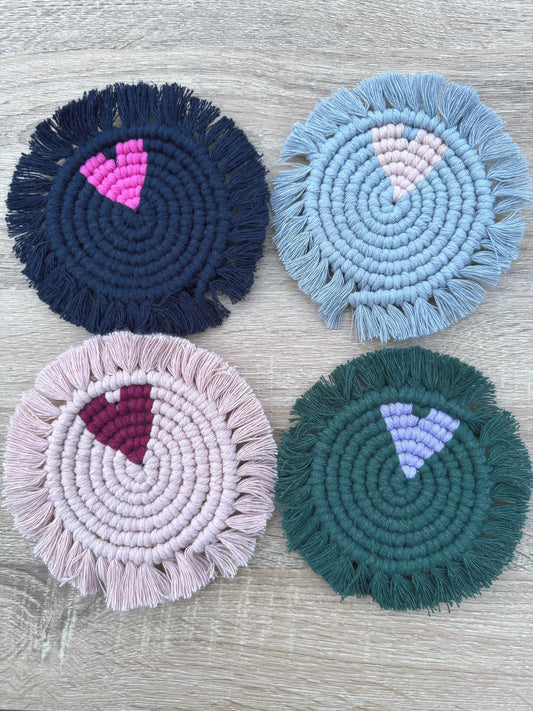 Four round macrame coasters with heart features and fringes in different colors on a wooden surface.