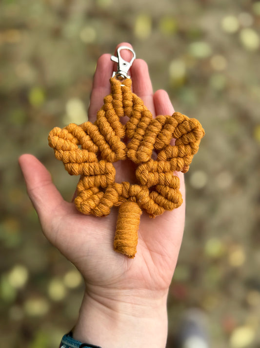 Hand holding a mustard yellow macrame maple leaf keyring against a blurred natural background