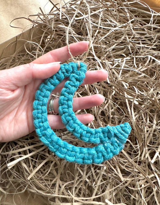 Hand holding a blue macrame crescent moon ornament on a bed of straw.