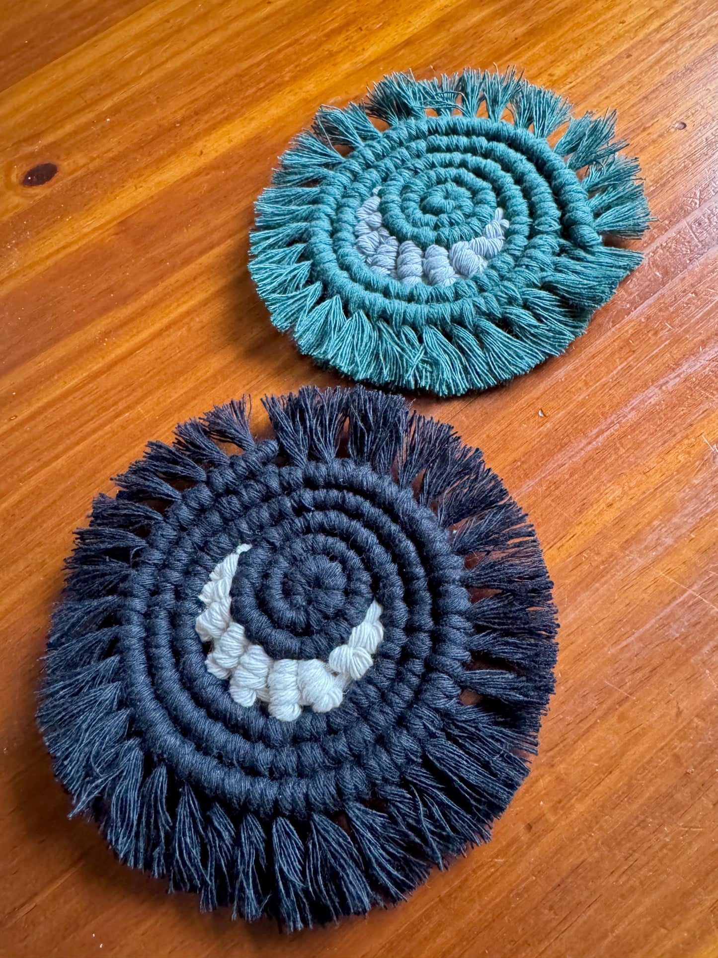 Two macrame coasters, one green and one black , on a wooden surface.
