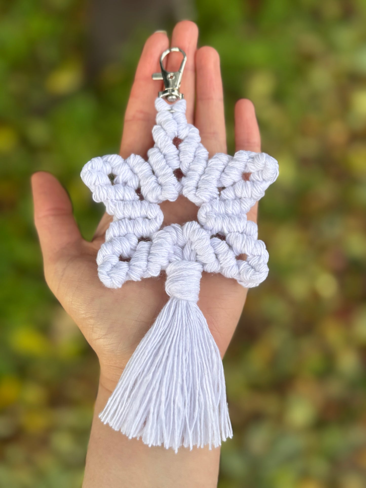 White macrame star keychain held in a hand with a blurred green background