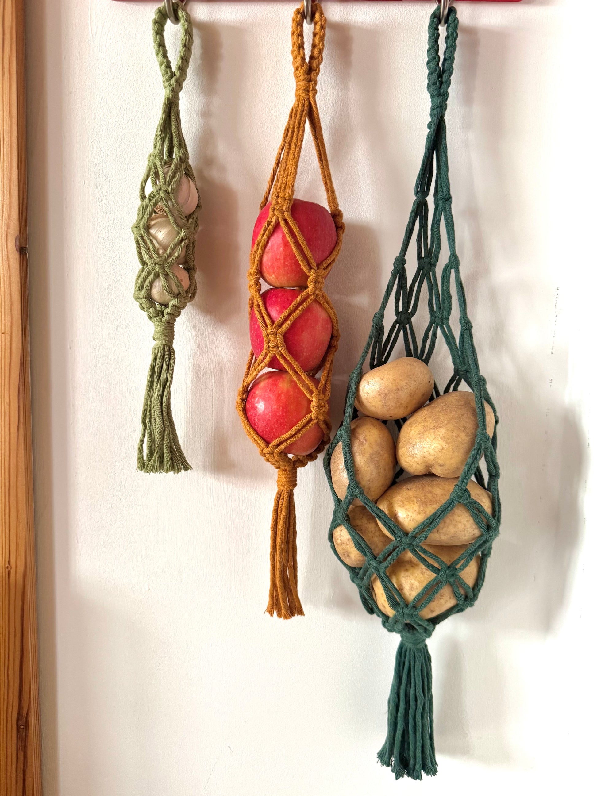 Three different coloured macrame net hangers with tassels holding garlic, apples and potatoes on a white wall.