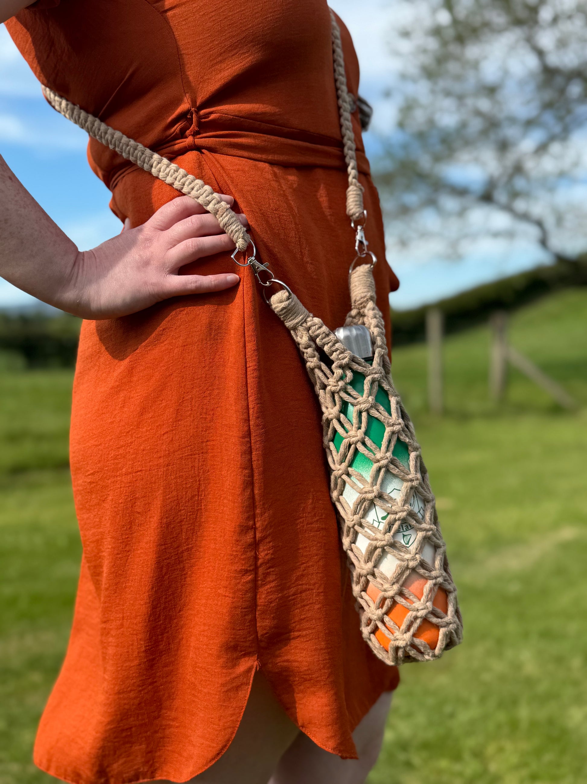 Person wearing an orange dress holding a macrame bottle holder outdoors.