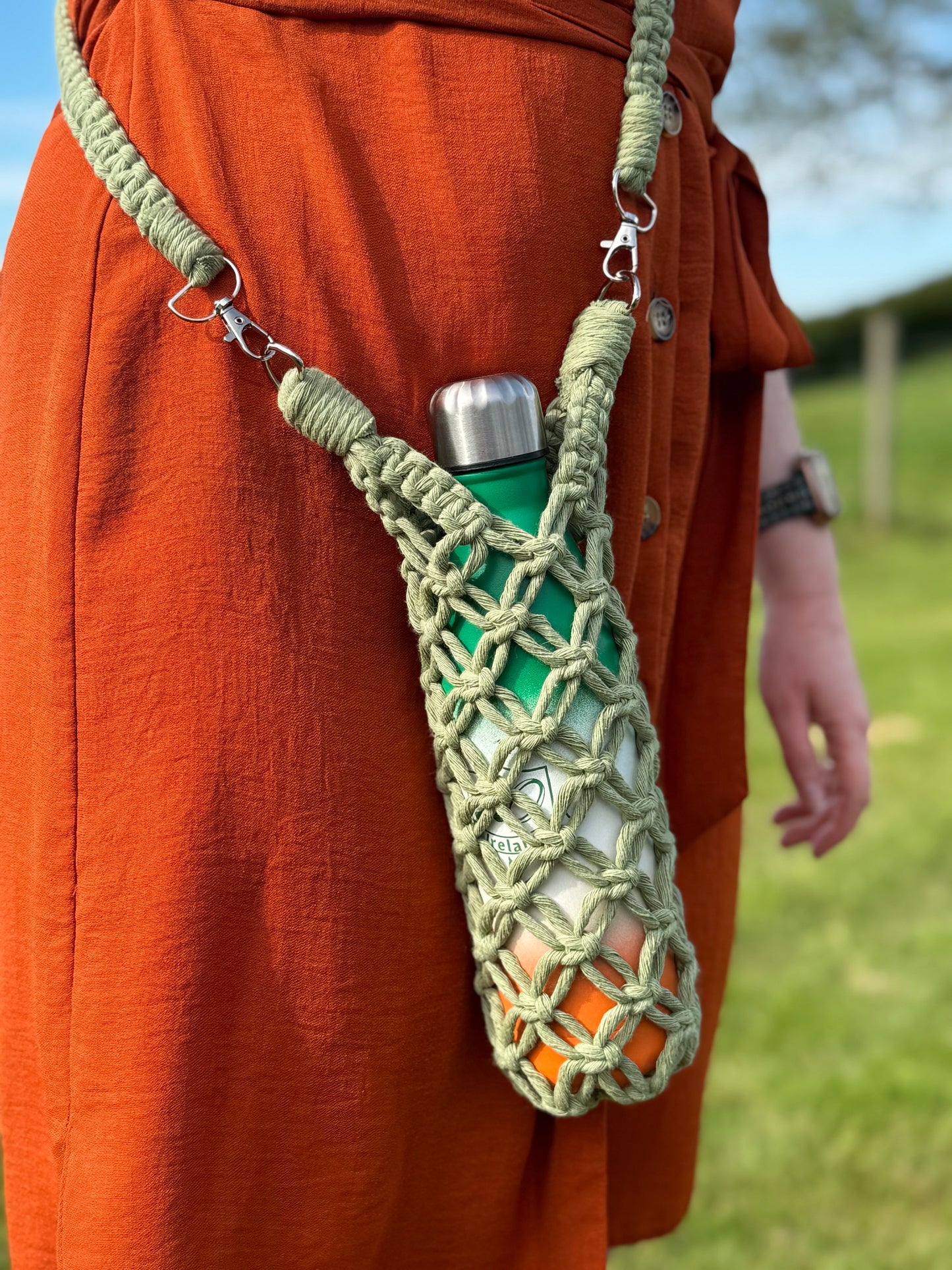 Person wearing a green water bottle with a macrame holder on an orange outfit outdoors.