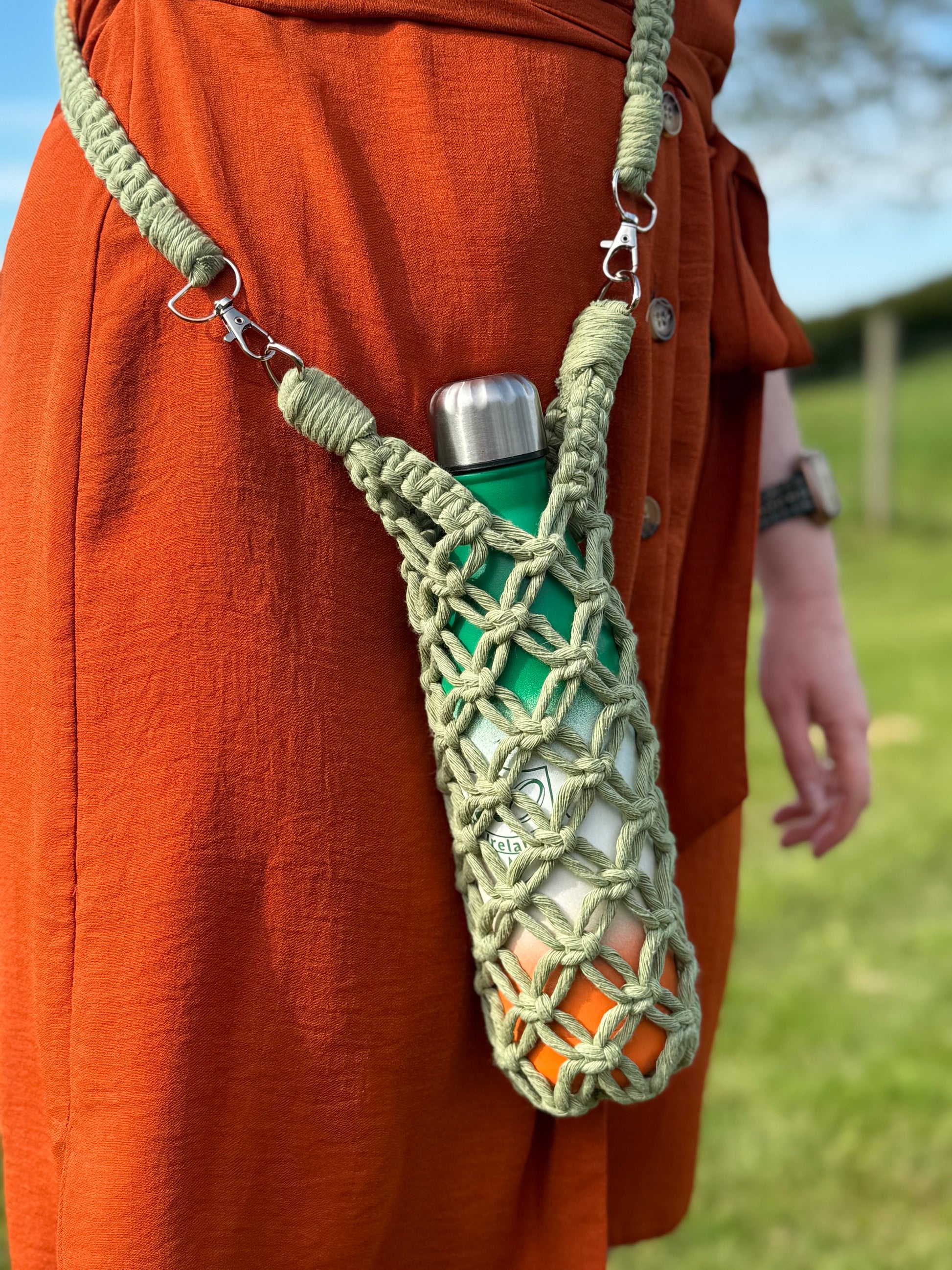 Person wearing a green water bottle with a macrame holder on an orange outfit outdoors.